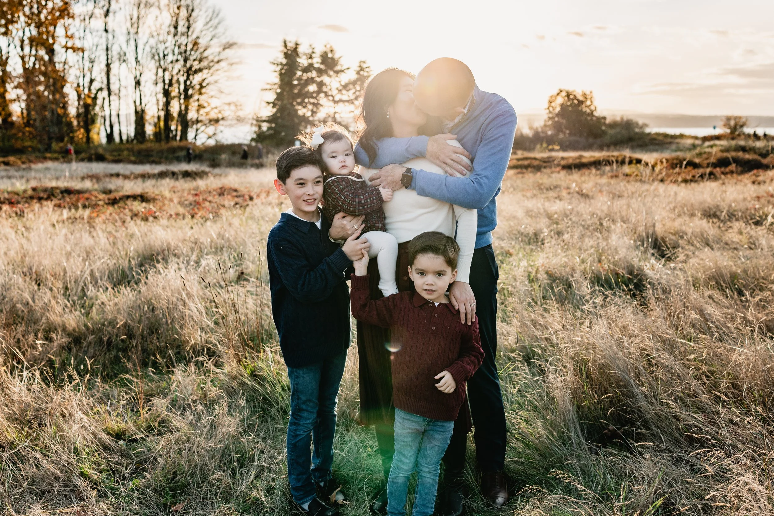 A candid moment where parents kiss while their two sons look at them and the mother holds the baby. Part of a fall Seattle family photography session in a meadow.