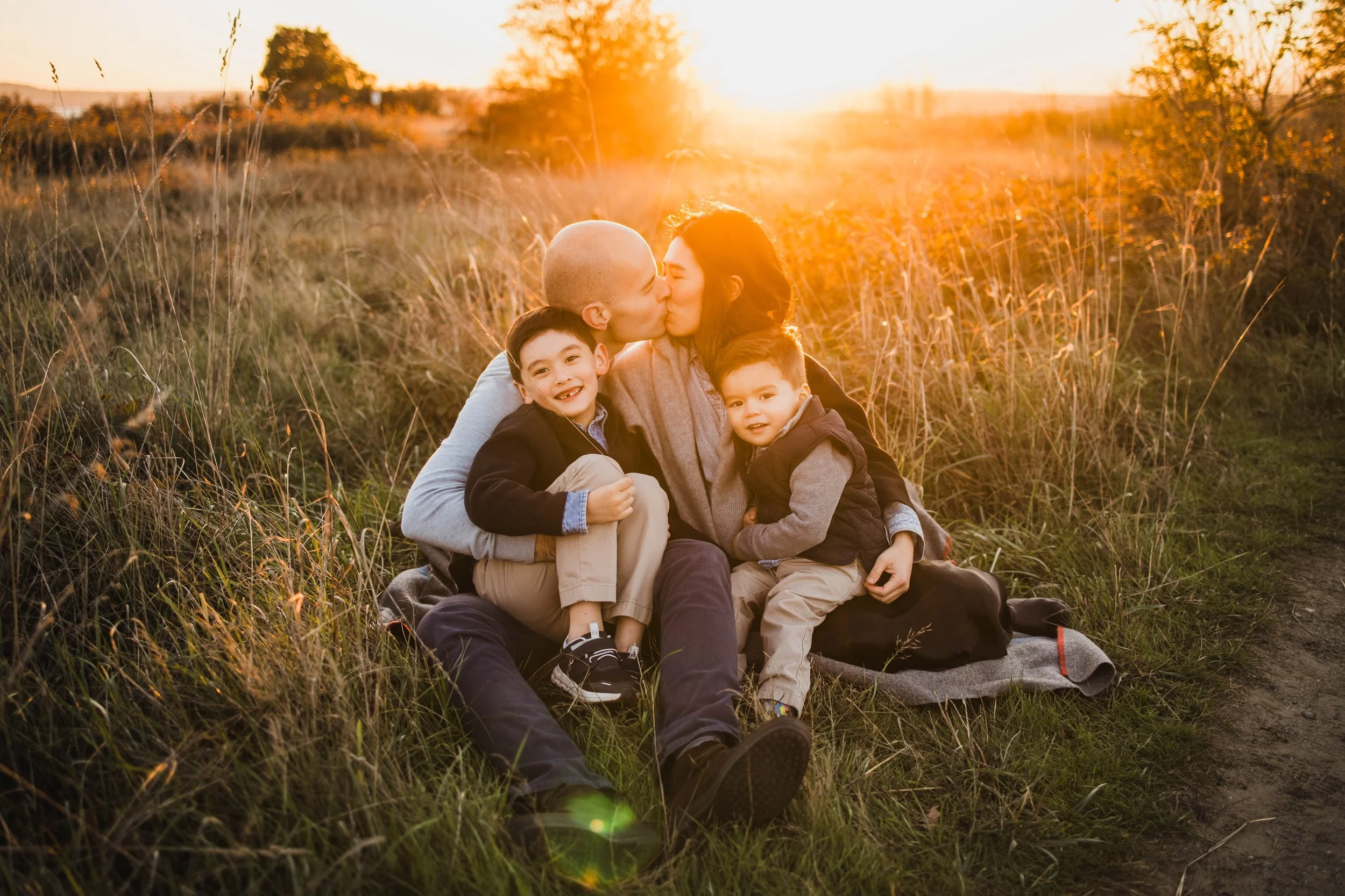 Parents share a kiss while sitting on a blanket in the tall grass at Discovery Park, with their two sons smiling in the foreground. Professional Seattle family photography at sunset.