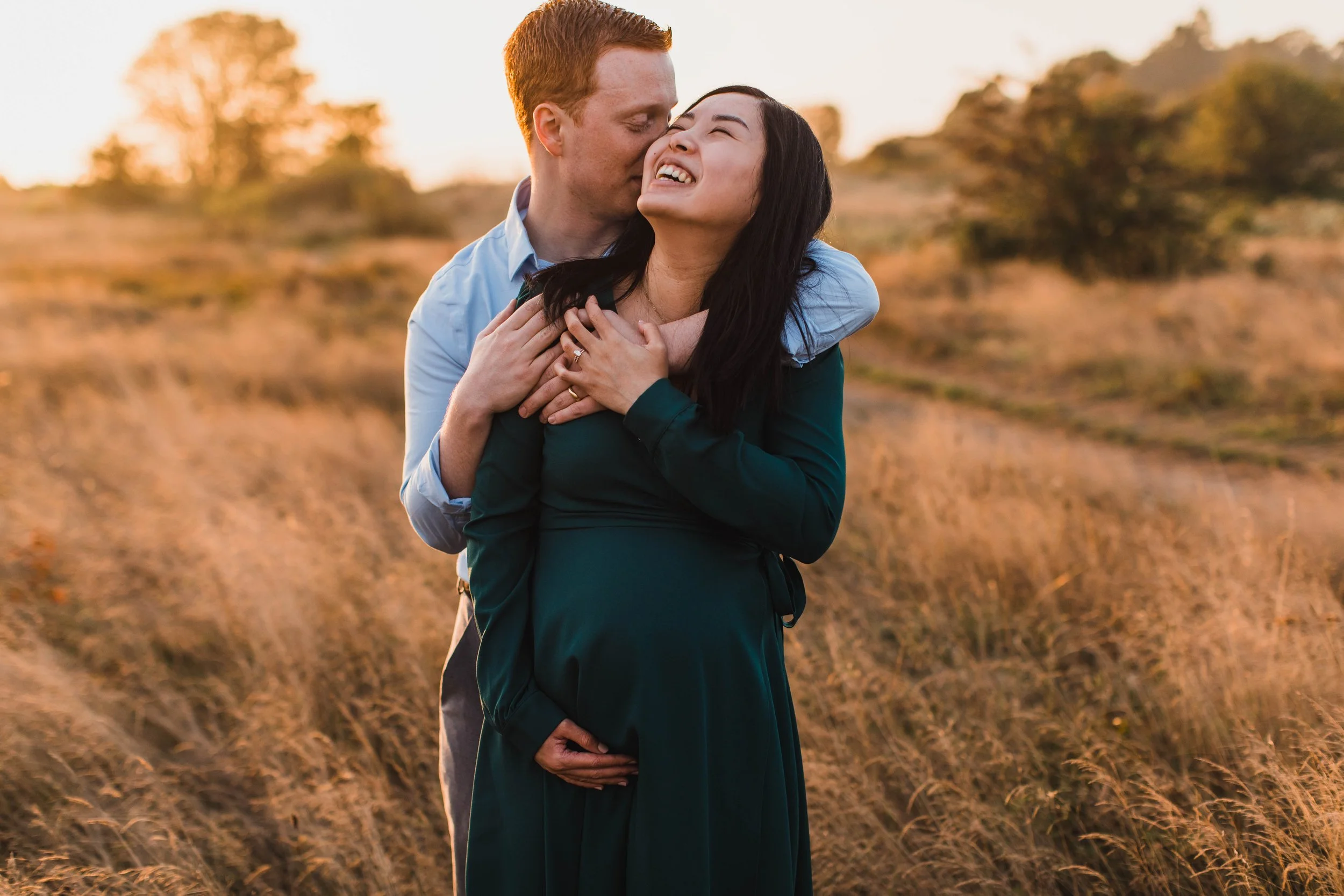 A man embraces his pregnant partner from behind as she laughs during a joyful seattle maternity photography session at sunset.