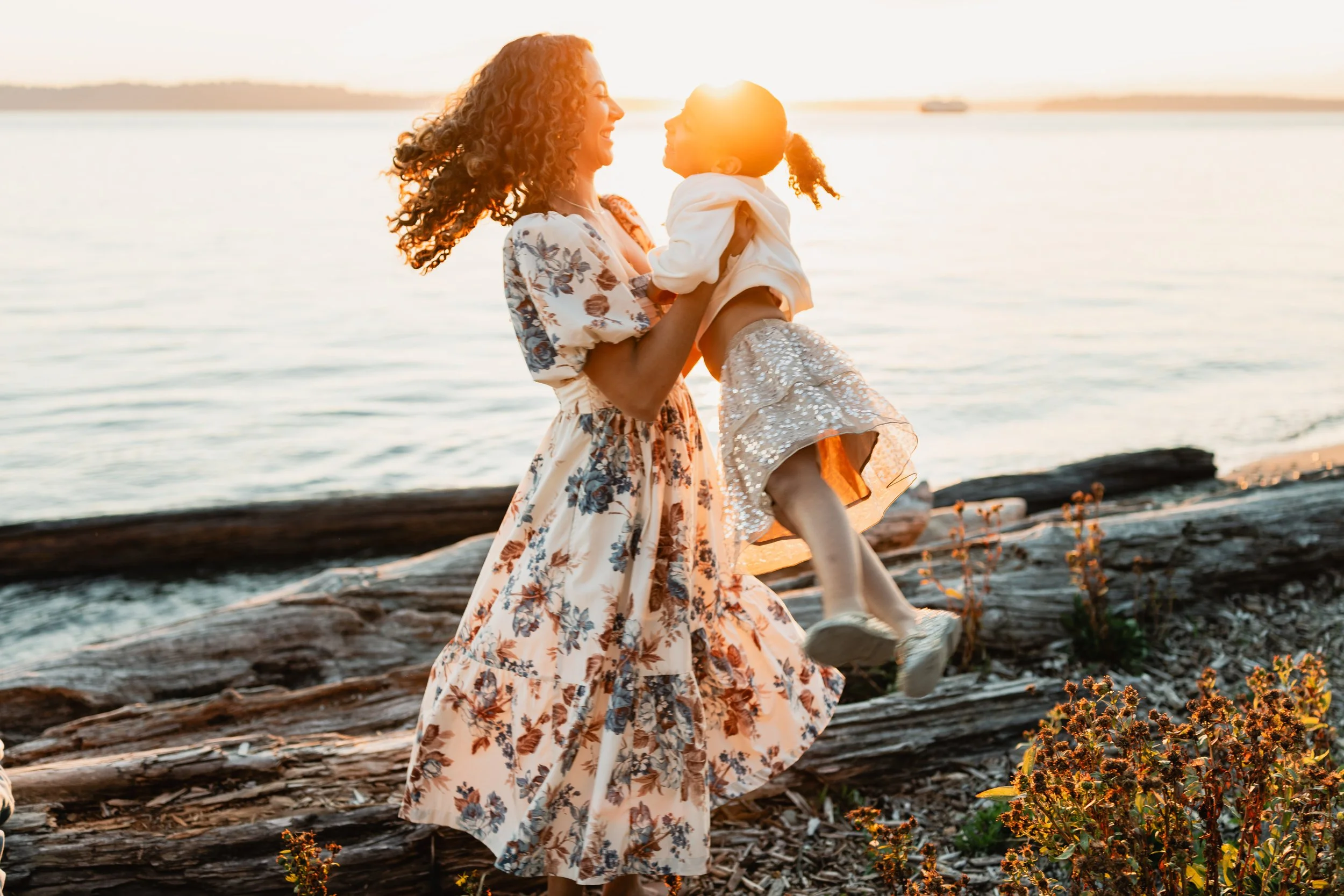 A mother in a floral dress playfully spins her daughter on the driftwood-lined shore during a West Seattle family photography session at sunset.