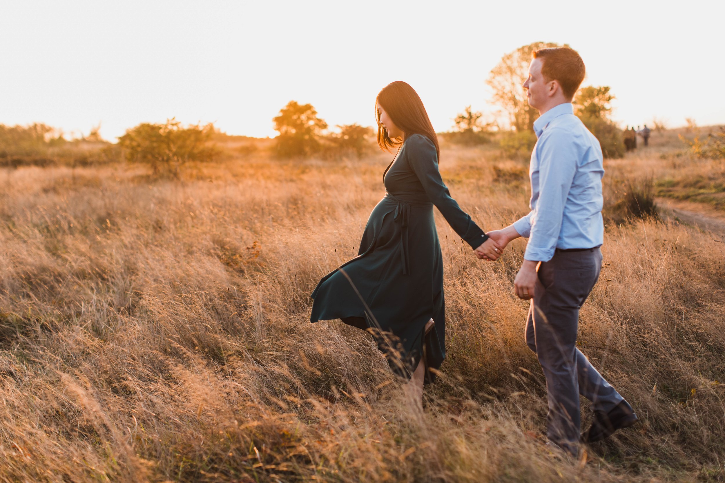 A couple holds hands and walks through a golden meadow during a sunset session for seattle maternity photography.