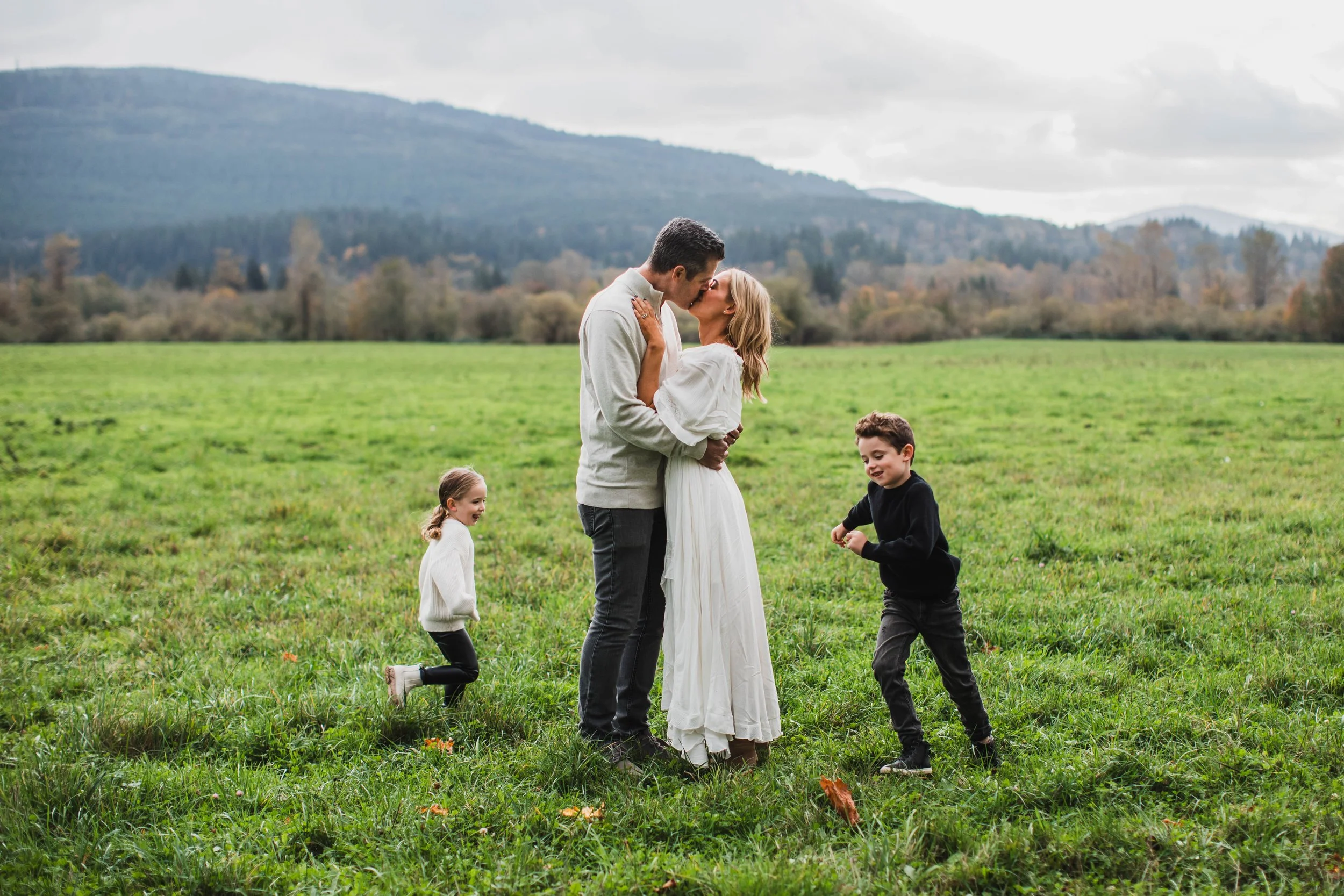 Outdoor photo shoot in North Bend for Seattle family photography. Parents kissing in the center as kids play, wide open field with mountain range view.