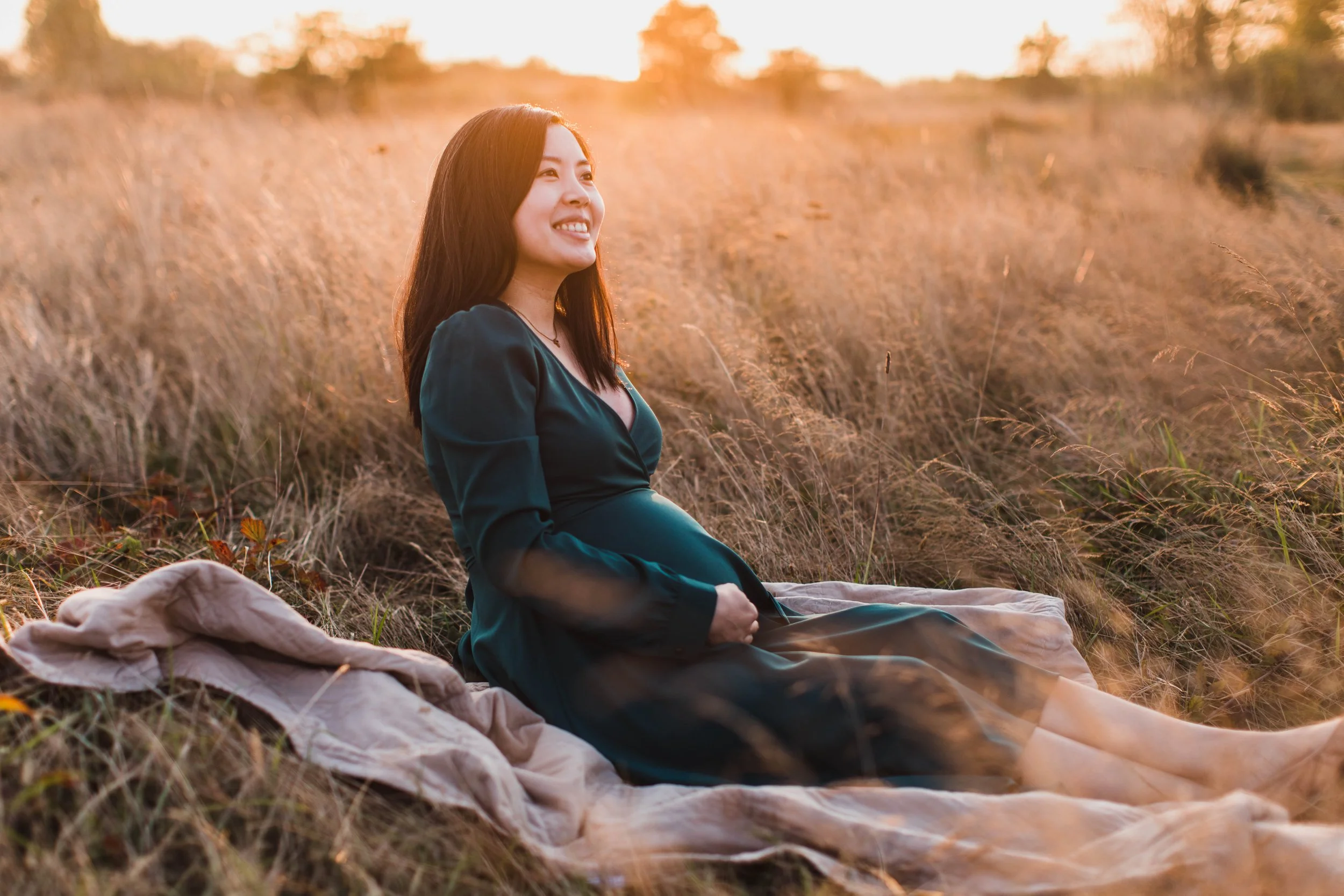 An expectant mother in a forest green dress sits in a golden field during a seattle maternity photography session at sunset.