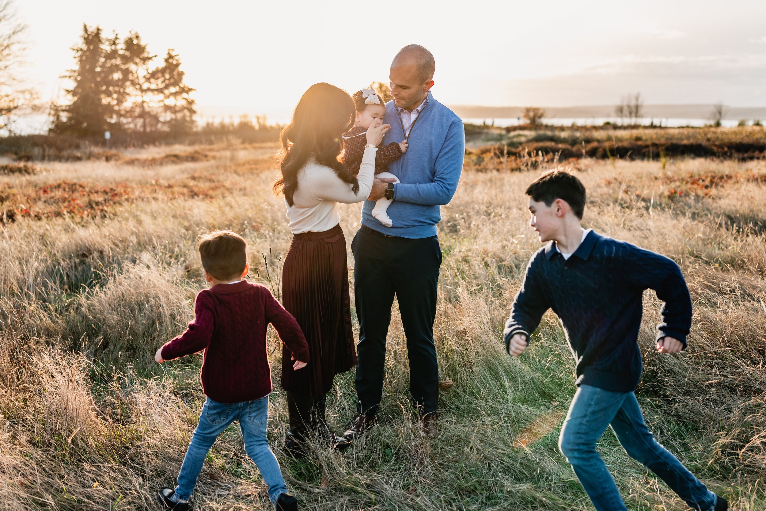 A candid moment where parents kiss while their two sons look at them and the mother holds the baby. Part of a fall Seattle family photography session in a meadow.