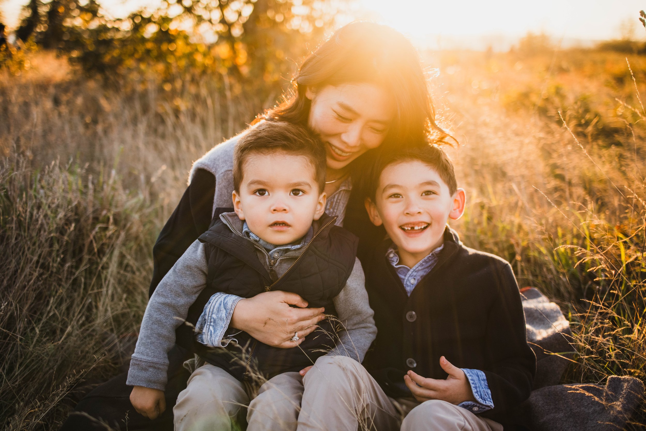 A mother sits in the tall, dry grass of Discovery Park with her two young sons, capturing a candid moment during a Seattle family photography session at golden hour.