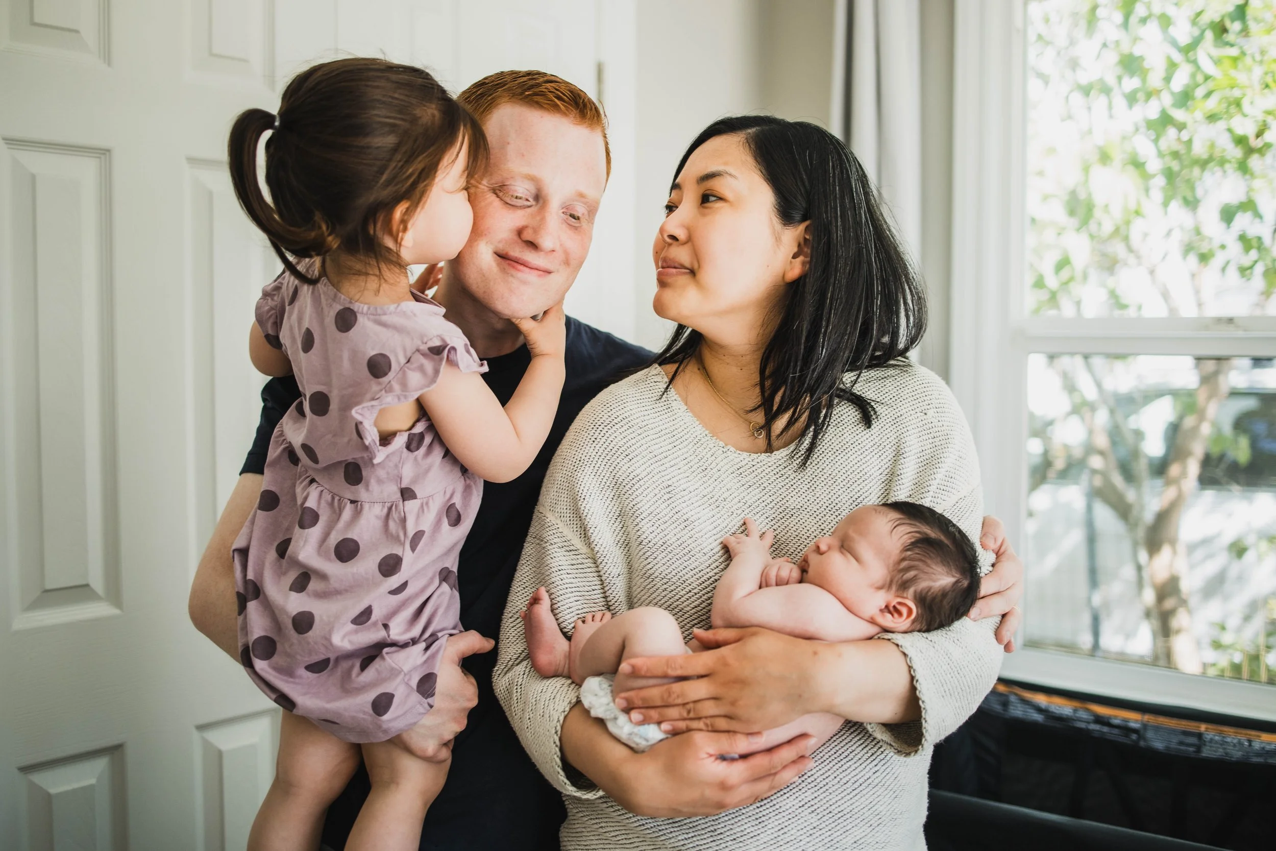 A candid in-home family portrait featuring parents and their toddler daughter kissing her father’s cheek, while the mother holds a sleeping baby. Part of a seattle newborn photography session.