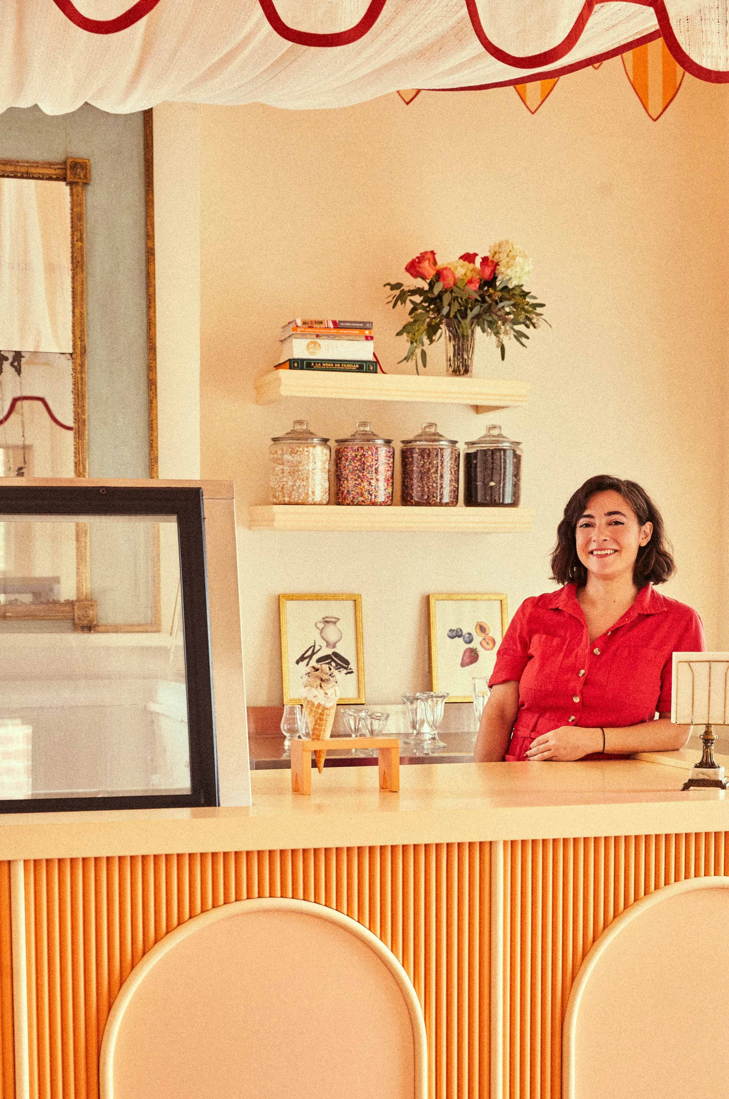 A woman in a red shirt smiling behind a wooden counter at a cafe or dessert shop, with jars of toppings, framed artwork, and a vase of flowers in the background.