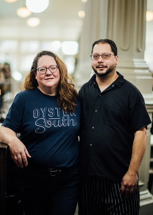 A woman and a man standing together indoors, smiling for the camera, with soft lighting and decorative columns in the background.