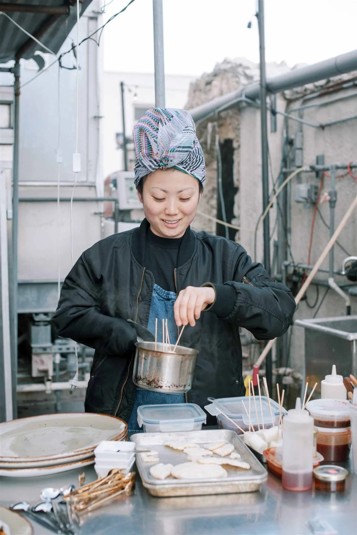A woman wearing a colorful headscarf and black jacket is cooking outdoors, handling skewers over a stove with various plates and containers of food around her.