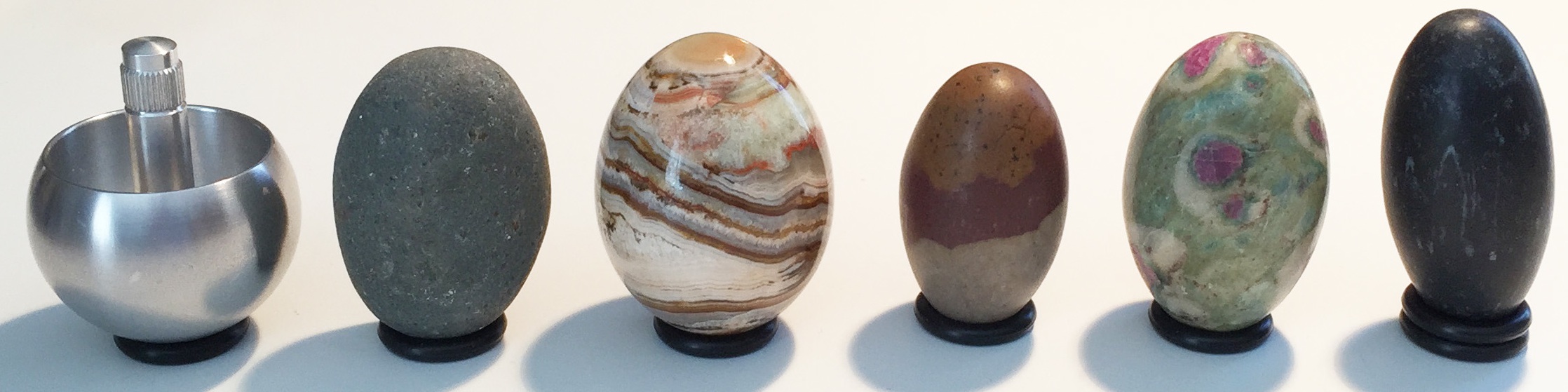 A variety of spinning objects: tippe-top; natural pebble; polished jasper stone egg; classic Lingam stone; ruby/fuschite Lingam stone; black Lingam stone. Each is about 5 cm tall.