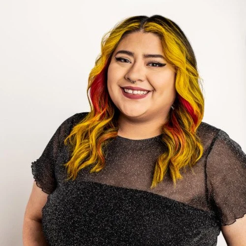 A woman with long, wavy hair dyed yellow and red, smiling and wearing a black, glittery top.