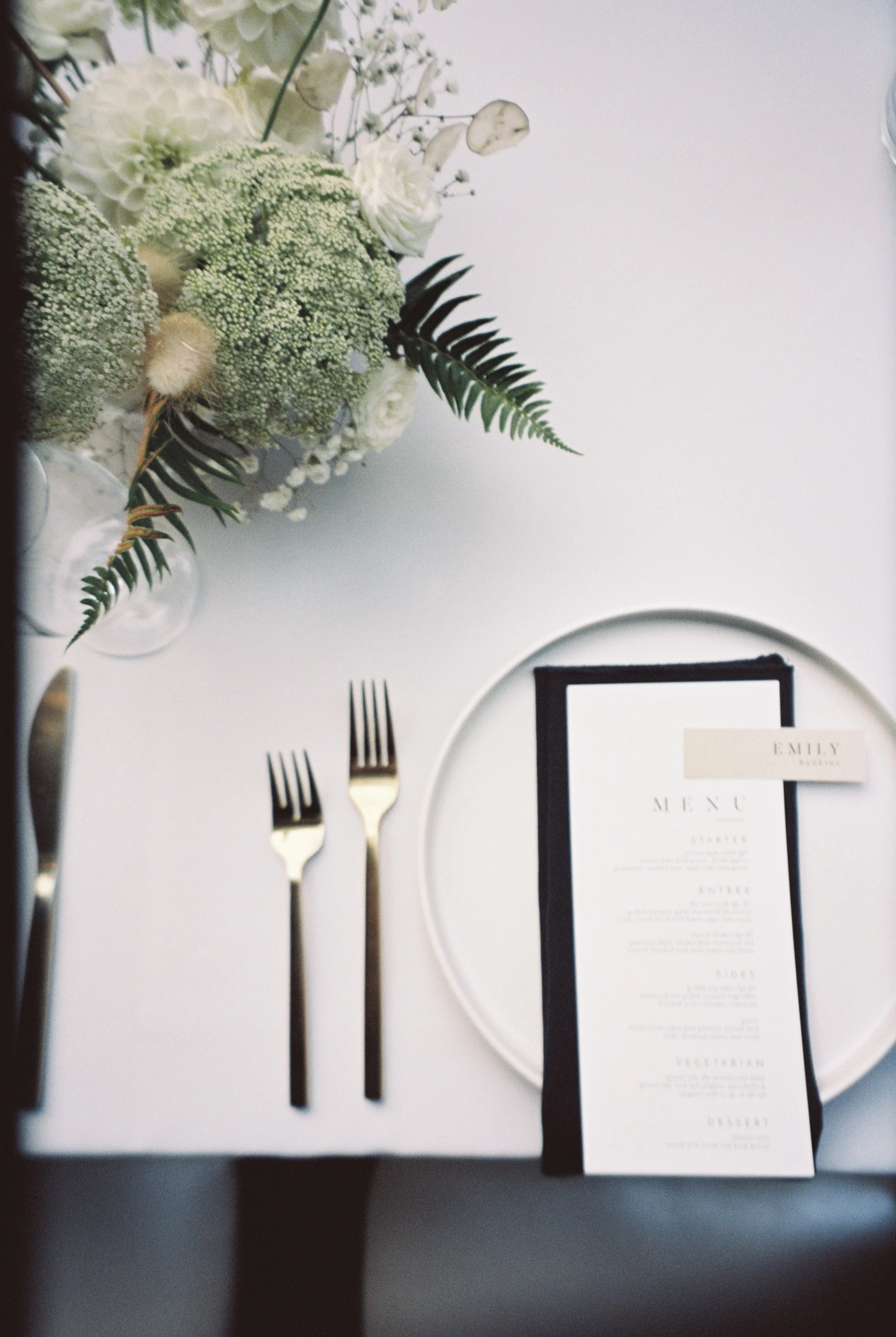 Elegant table setting with cutlery, a menu card, a white plate, and a floral centerpiece with white and green flowers and fern leaves.
