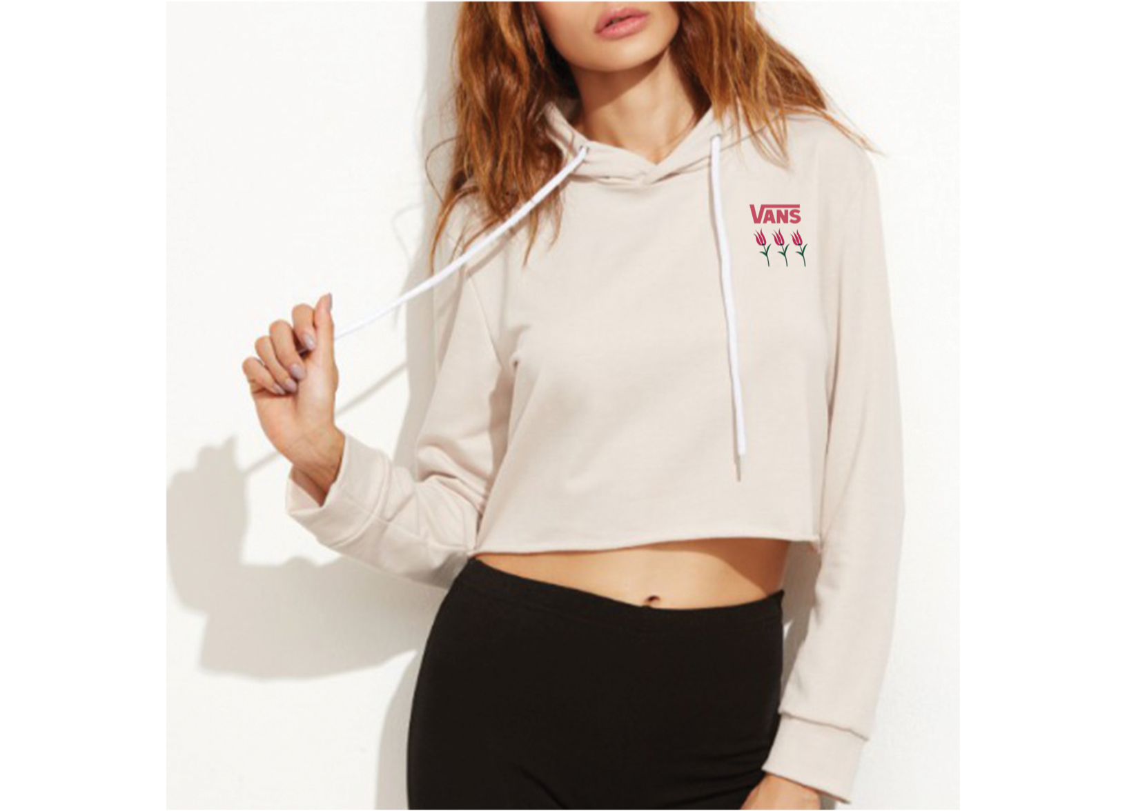 A woman wearing a cropped beige hoodie with a small pink Vans logo and three pink tulip flowers on the left chest, holding the drawstring of the hoodie. She is also wearing black pants against a white background.