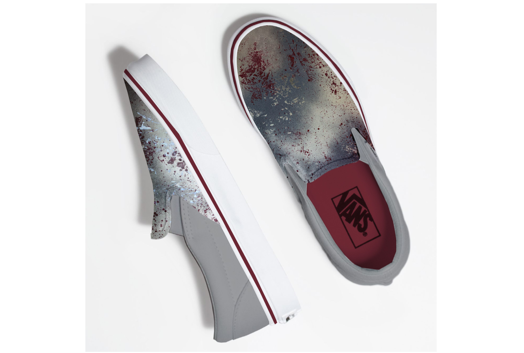 A pair of Vans slip-on sneakers with a gray and red splatter pattern design, placed on a white surface, showing the top and side views.