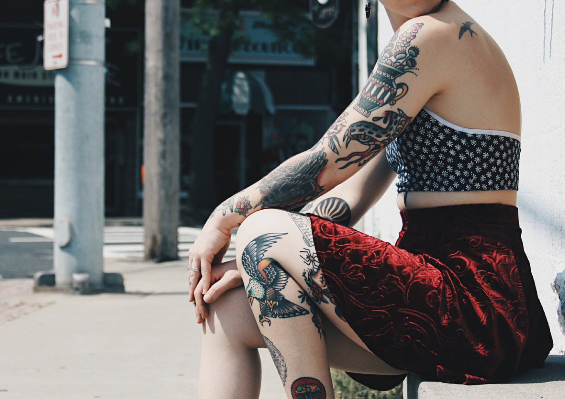 A person with colorful tattoos sitting on a sidewalk against a white wall, wearing a black and white floral crop top and red shorts with a black pattern. Their tattoos include a snake on their arm, a bird on their thigh, and a dragon on their leg.