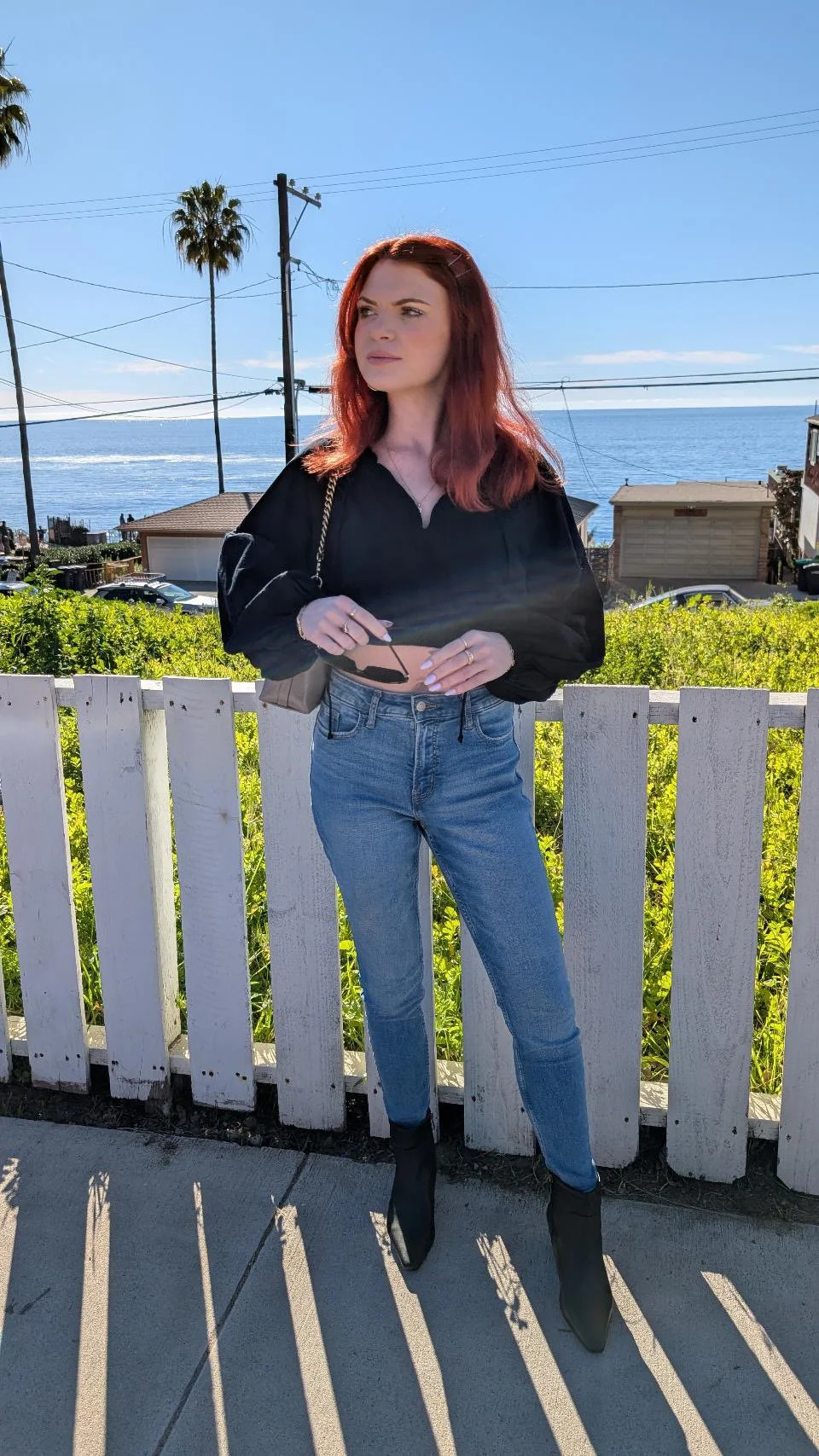 Young woman with red hair wearing black top, light blue jeans, and black boots standing outdoors near a white picket fence, holding a phone, with a scenic view of the ocean, palm trees, and a sunny blue sky in the background.