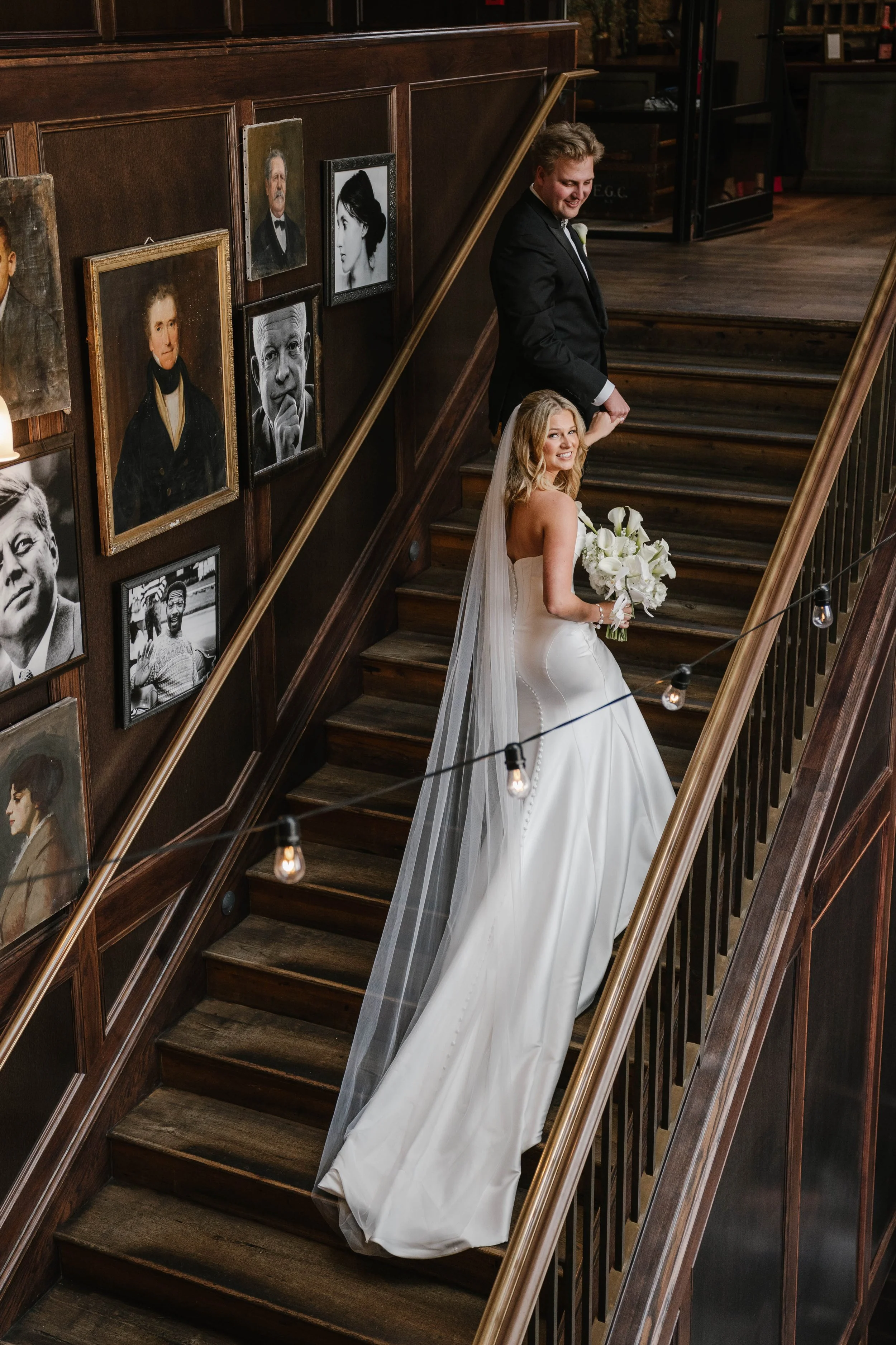 A bride and groom holding hands on a wooden staircase in a vintage-style venue decorated with framed portrait photos on the wall.