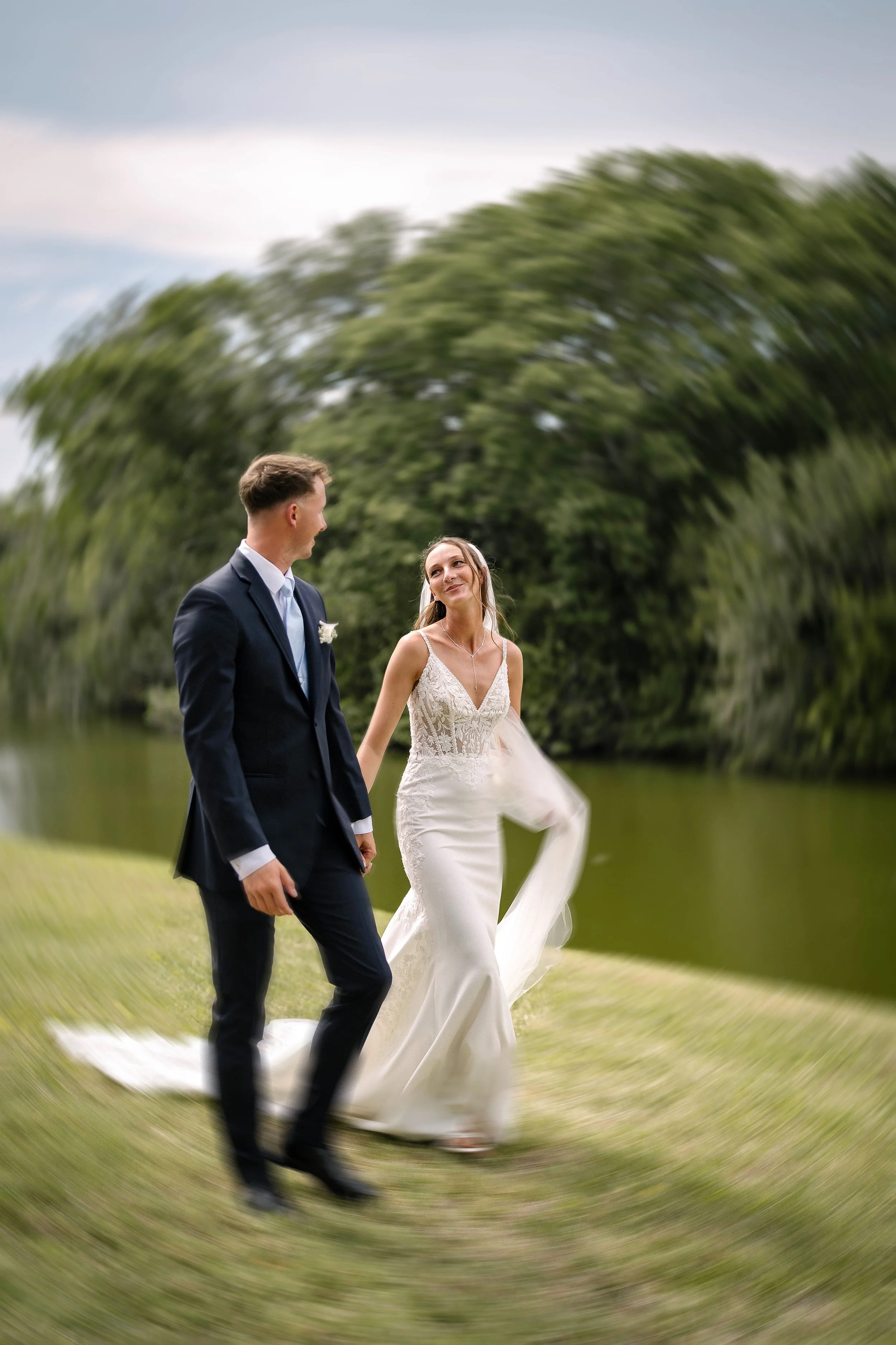 A bride and groom holding hands and walking outdoors on a grassy area, with trees in the background. The bride is looking at the groom, who is smiling and looking ahead. The bride is wearing a white lace wedding dress, and the groom is in a dark suit.