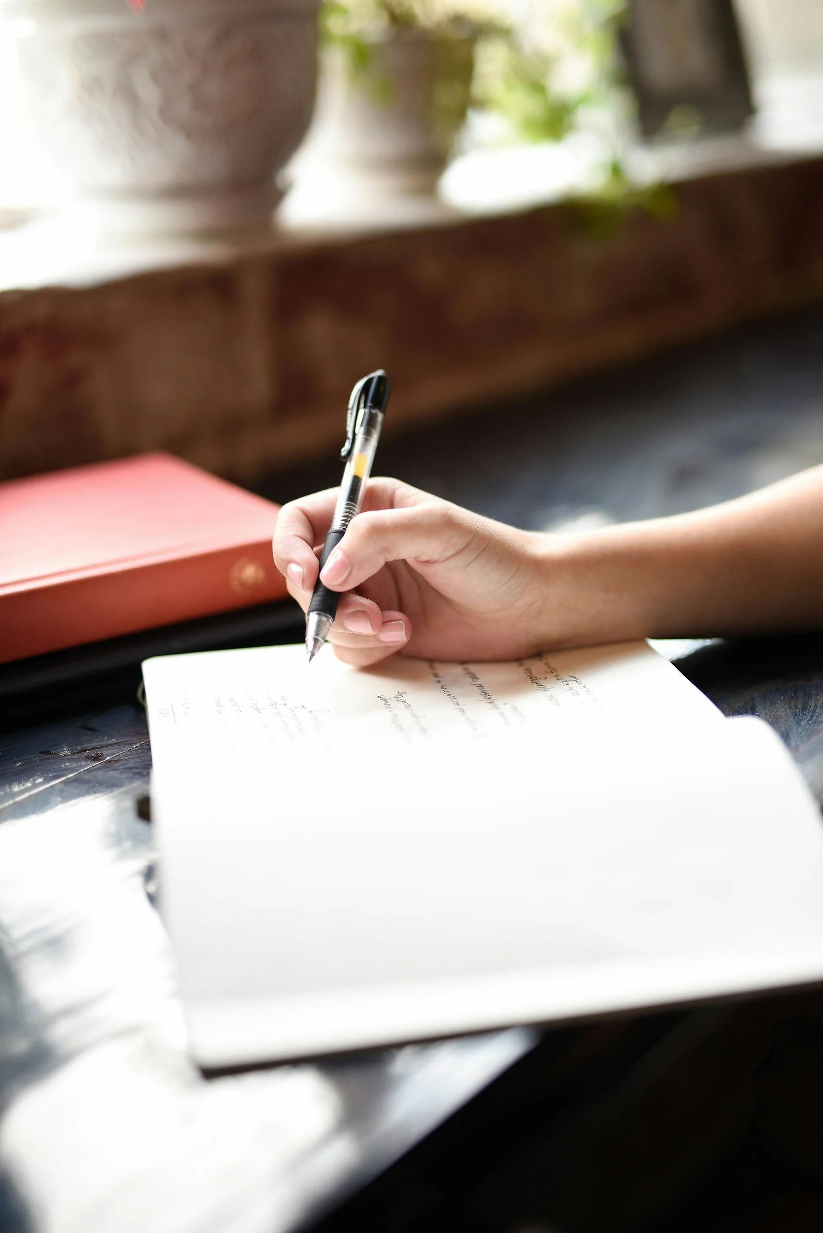Women's hand writing in a journal on a black desk with red book in the background