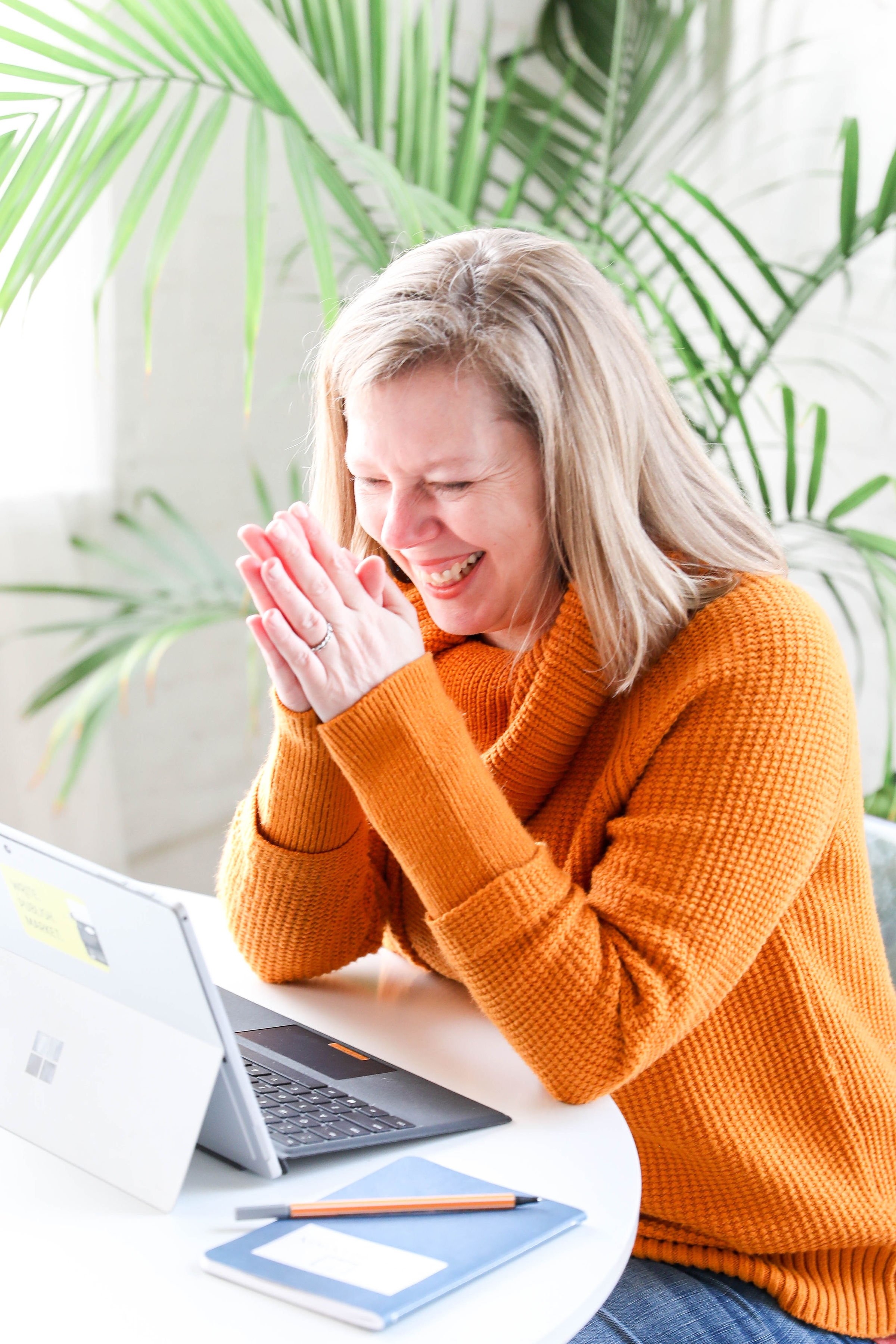 Jodi Brandon wearing orange sweater with hands folded and laughing in front of her open silver laptop