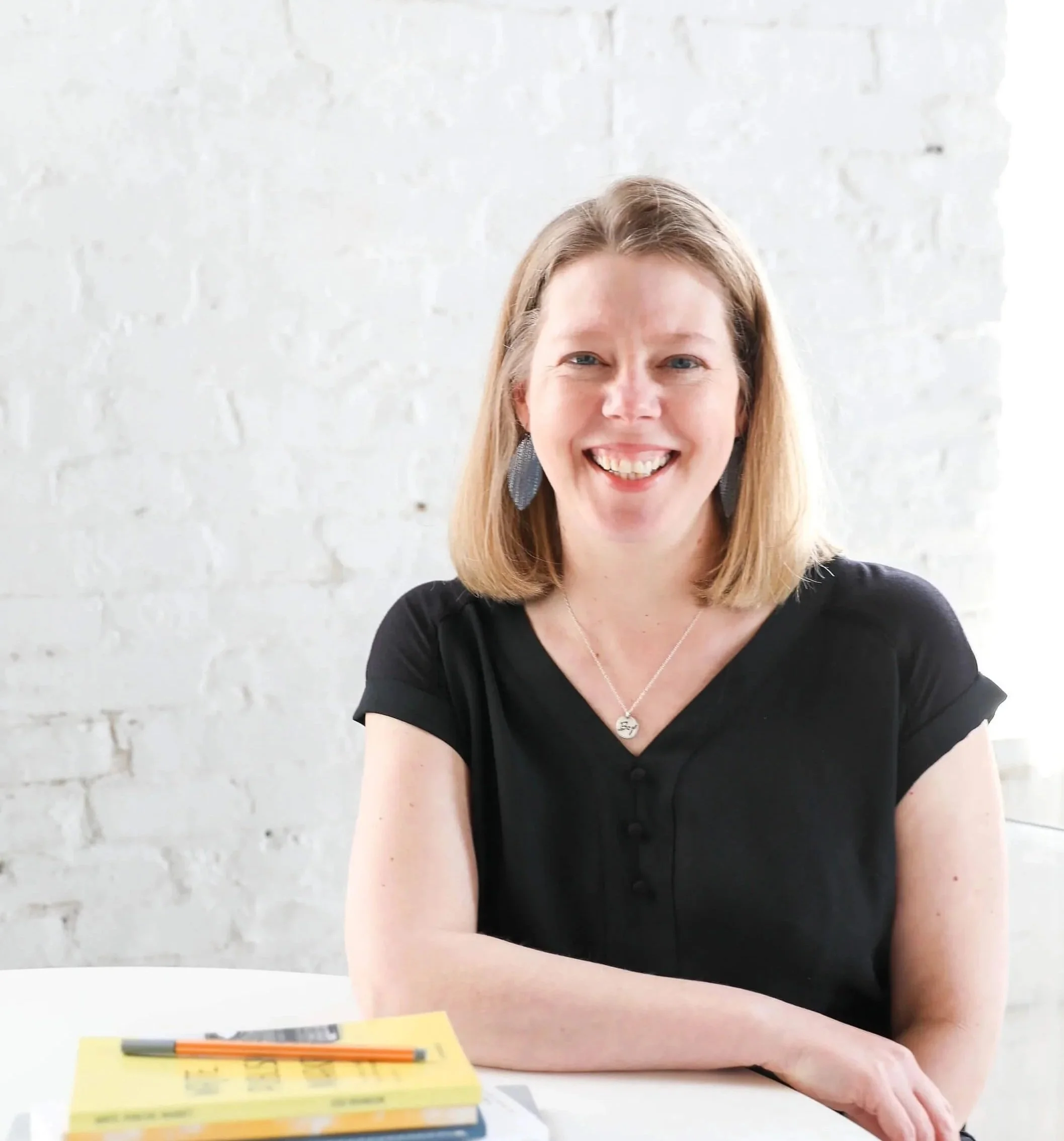 Jodi Brandon in short sleeve black shirt sitting at white table with stack of books smiling