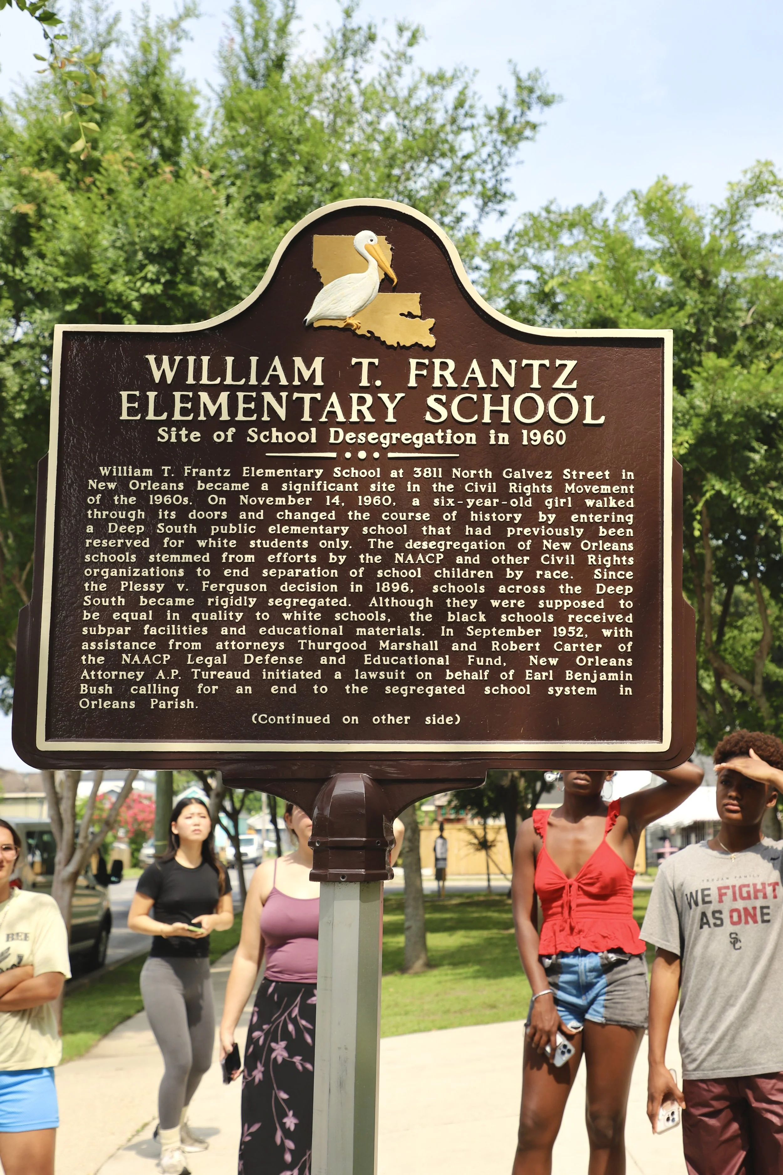  Students stand surrounding a sign posted in front of a school. This sign explains the event that prompted the desegregation of schools.  