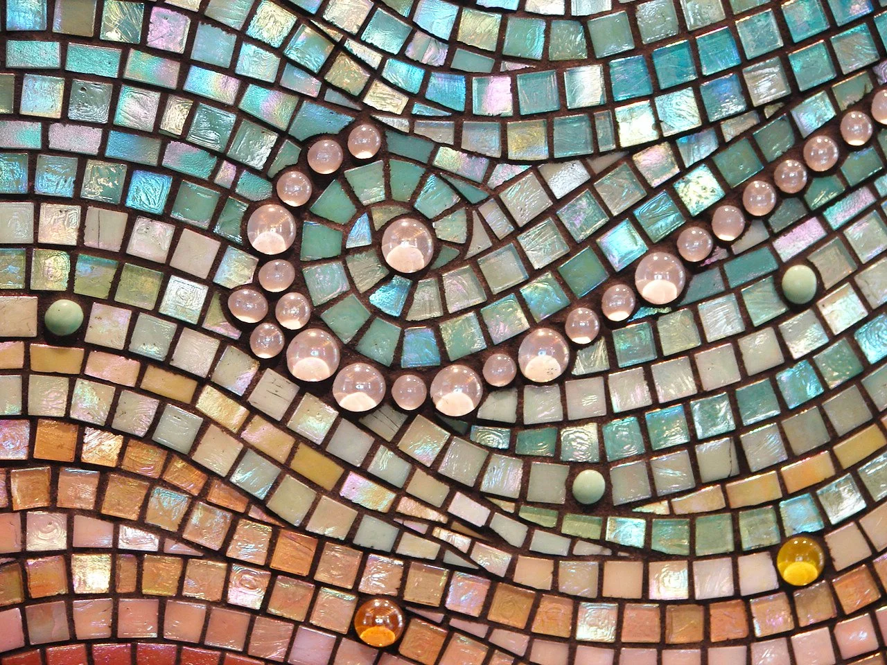 Close-up detail of Mokuluas Islands mosaic backsplash showing iridescent glass gems, pebbles, and spiral sand patterns in warm peach and green tones