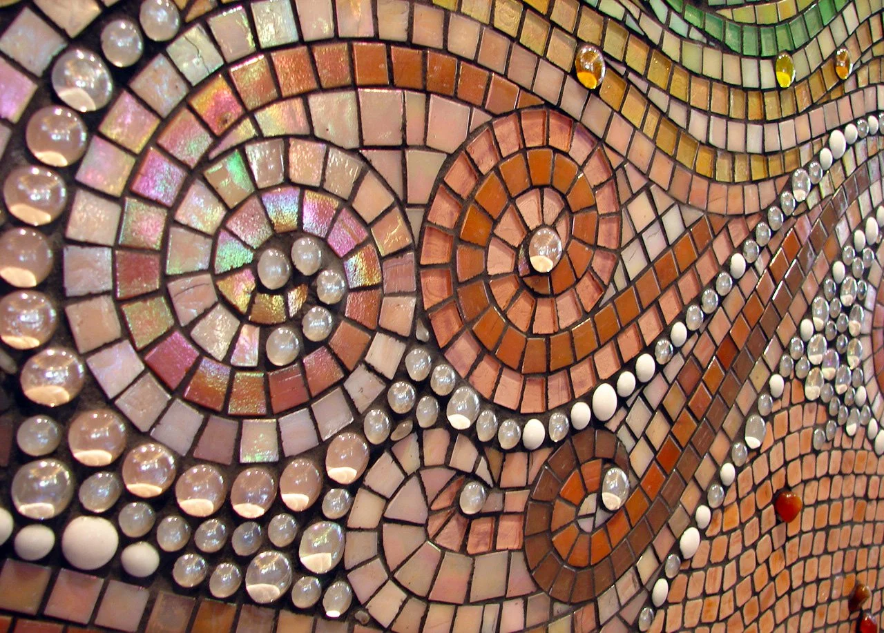 Close-up detail of Mokuluas Islands mosaic backsplash showing iridescent glass gems, pebbles, and spiral sand patterns in warm peach tones