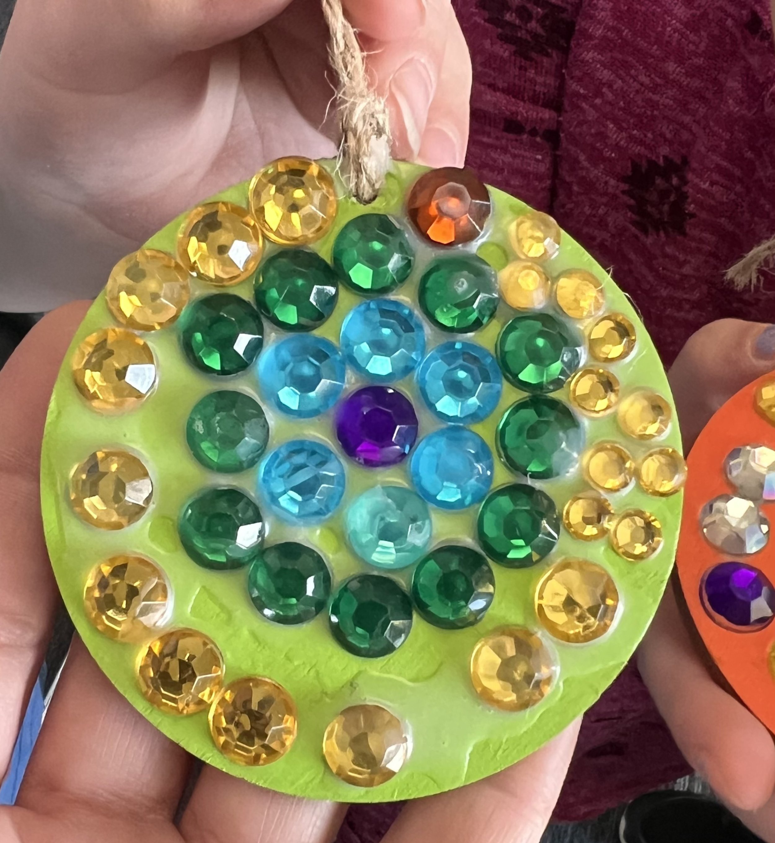 Children's hands holding two completed gem mosaic medallions made during the Cycles public art commission student participation project at Ida Nason Aronica Elementary School
