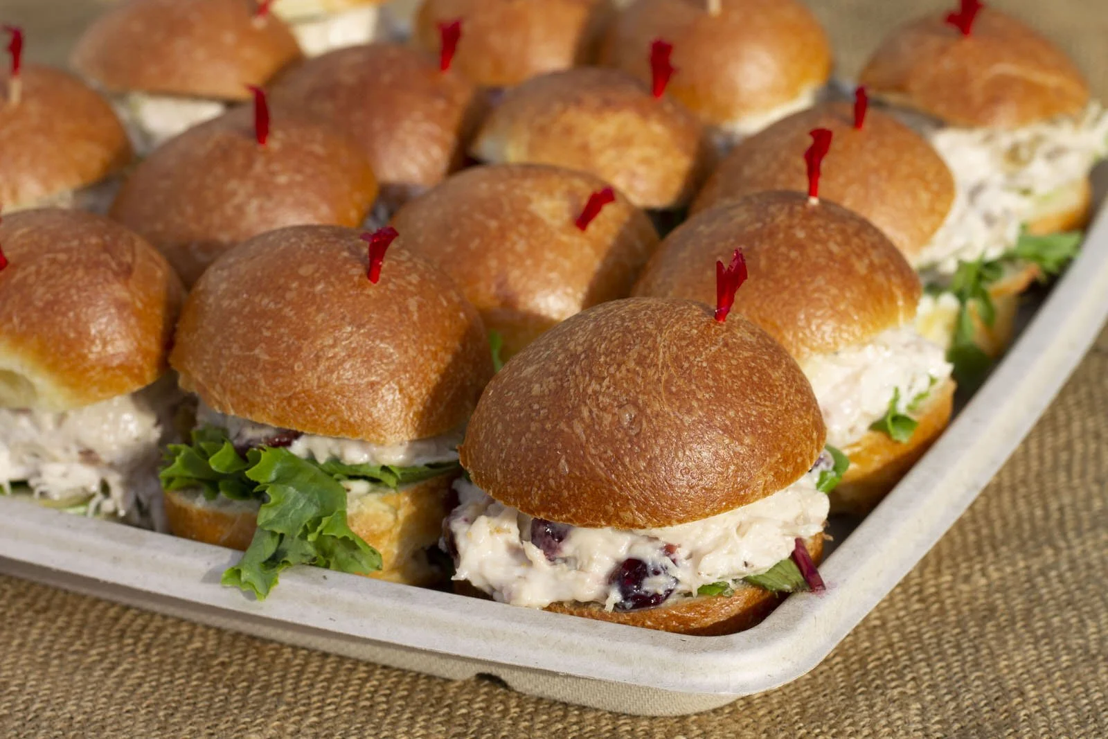 Tray of small sandwiches made with slider buns, leafy greens, and creamy filling, secured with red toothpicks.