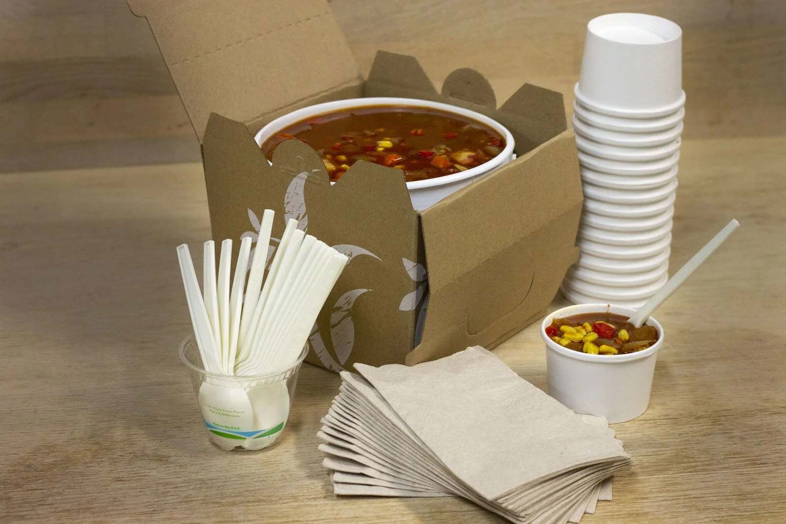 A large cup of chili in a cardboard to-go container with a stack of white disposable cups beside it, a small cup of chili with a spoon, a pile of white foam food containers, and a pile of napkins on a wooden table.