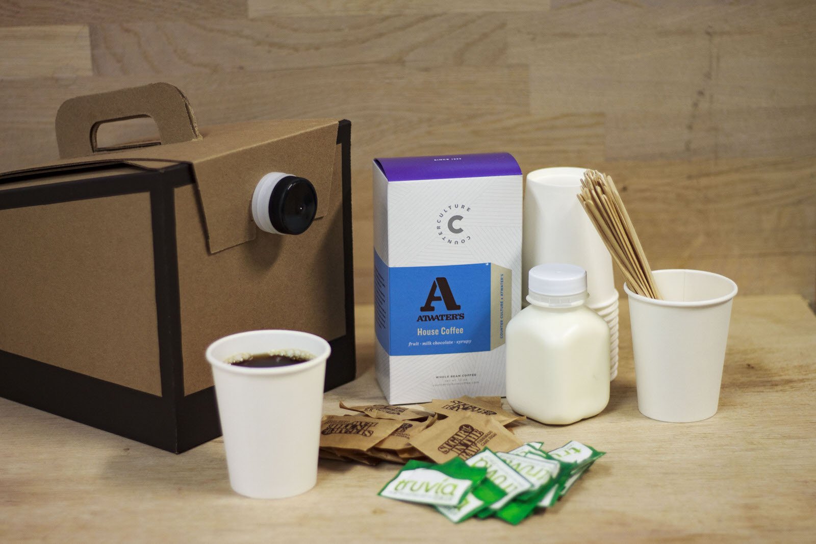 A coffee drink is in a white paper cup in front of a cardboard coffee box, with coffee packs, green sugar packets, milk cartons, a stack of cups, and wooden stir sticks on a wooden table.