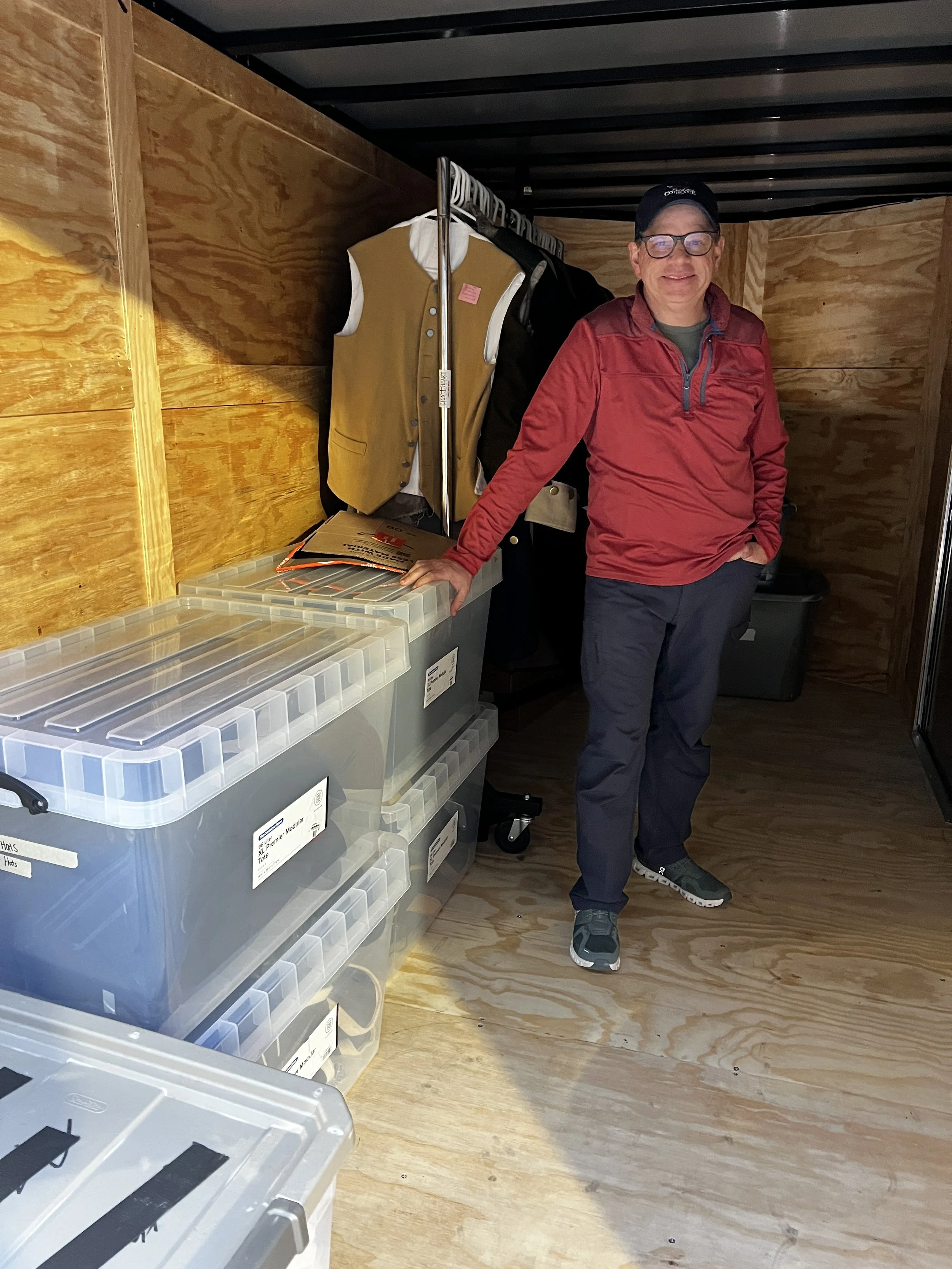 A smiling man in a red jacket and glasses standing in a small room with wooden walls and floor, next to plastic storage bins and a clothing rack with jackets and vests.
