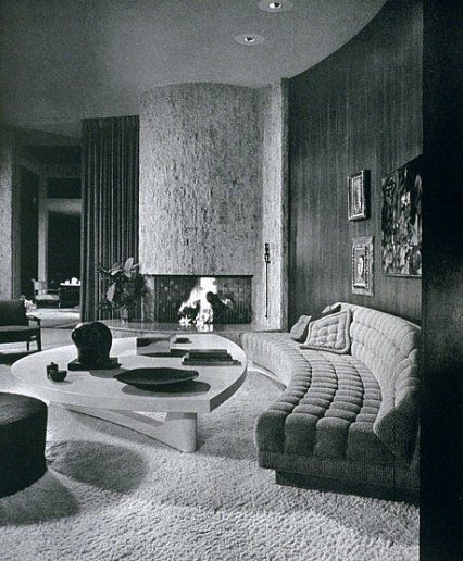 The living room of Mr. and Mrs. Eugene V. Klein’s Trousdale Estates, Beverly Hills, residence. The room features a walnut paneled wall, custom tufted curved sofa, coffee table of picked cork and a large travertine fireplace. Interior design by Arthur Elro