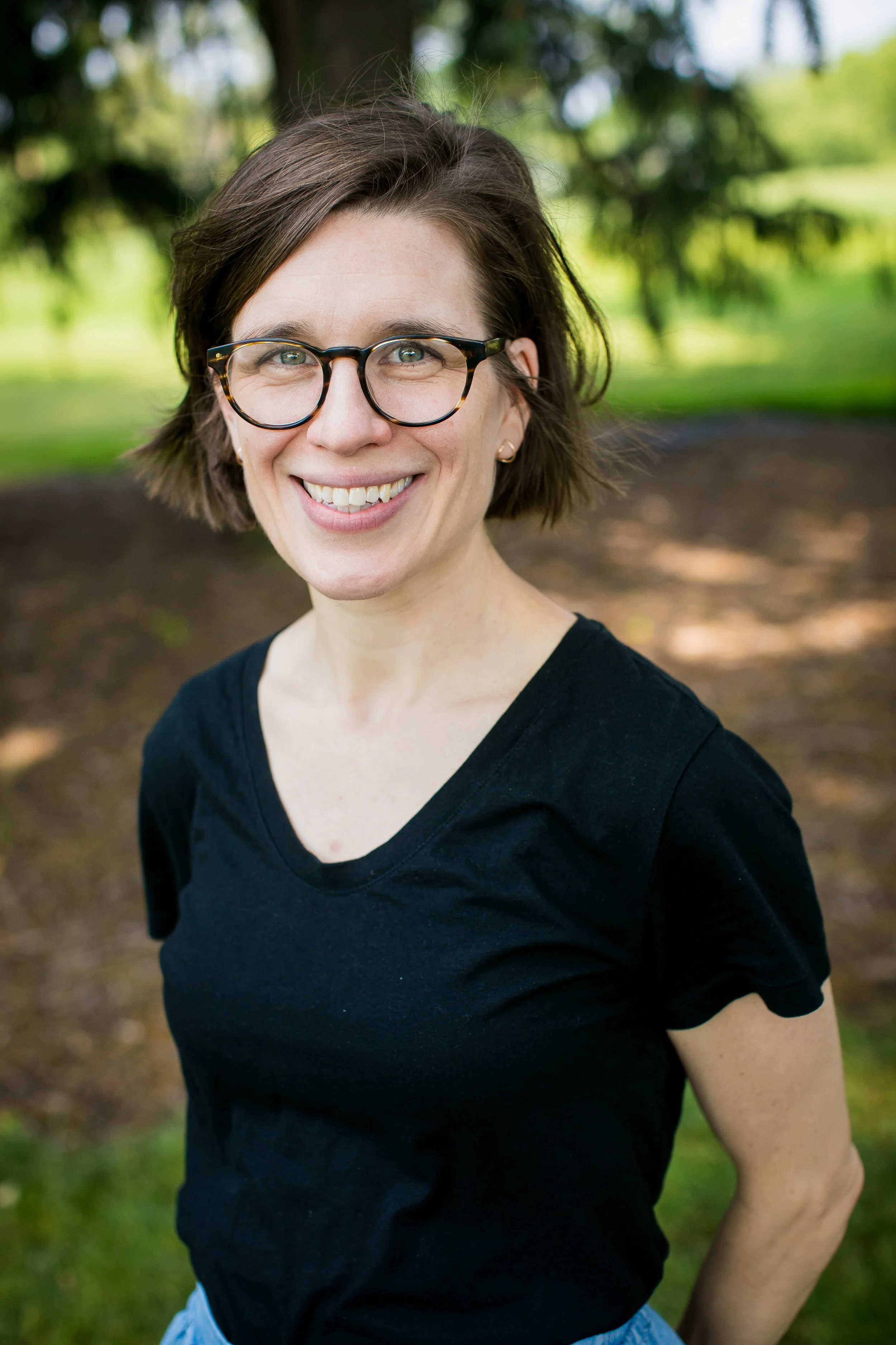 Headshot of a white woman in a black t-shirt. She is wearing glasses and standing outside, with dirt and grass in the background