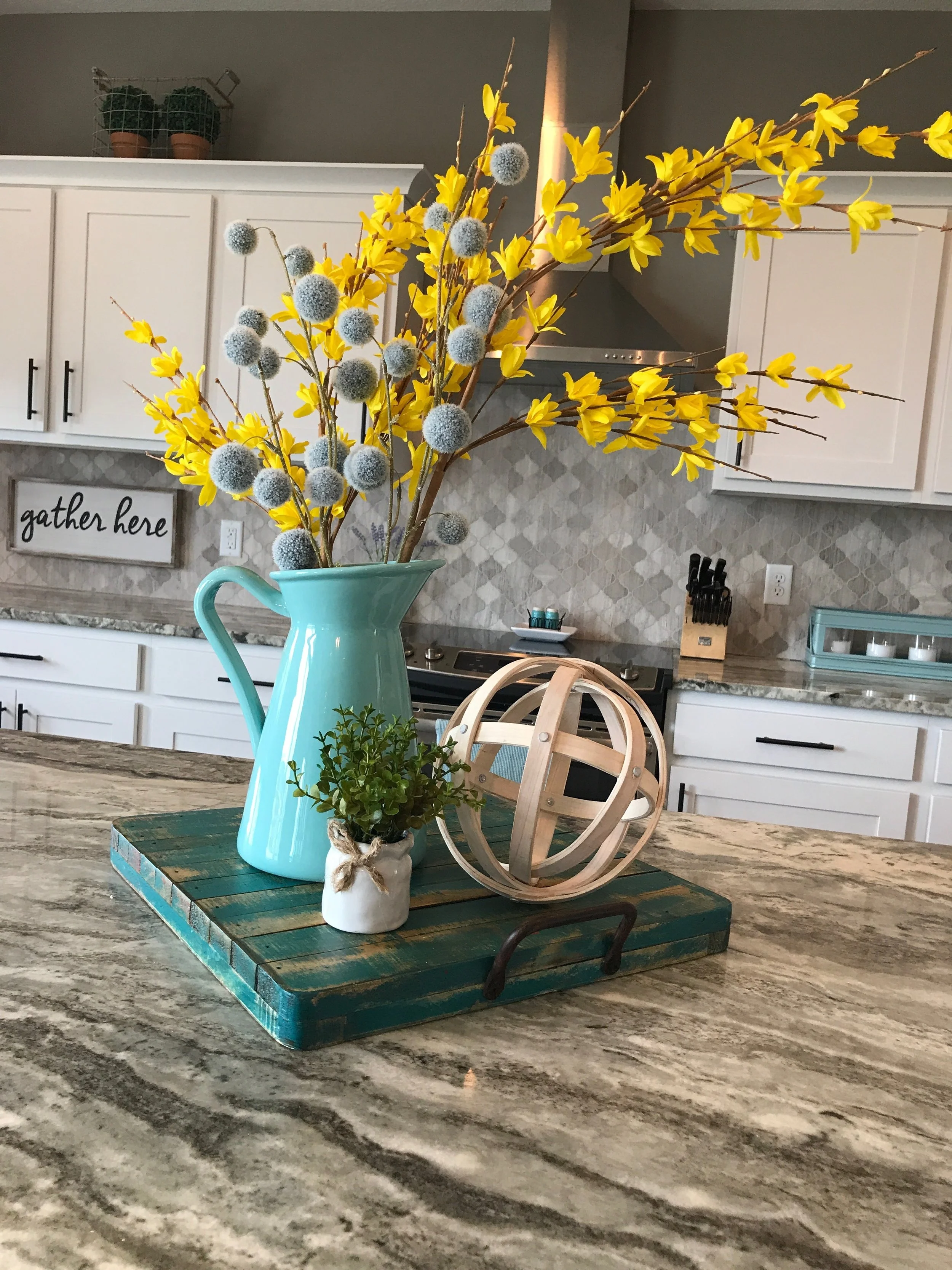 Kitchen vignette using a turquoise wooden tray, wood orb, small plant, a light turquoise pitcher filled with yellow floral stems, and blue billy buttons. RustopiaConsulting.com