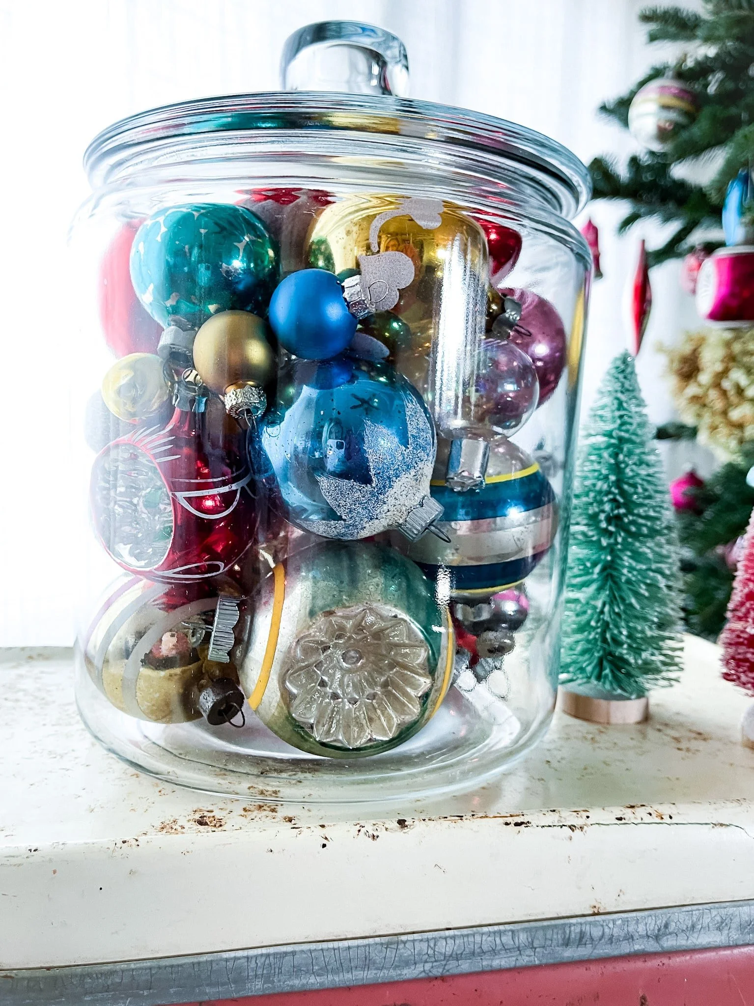 Vintage mercury glass and Shiny Brite Christmas ornaments in a glass canister, sitting on a vintage cooler and next to bottle brush trees.