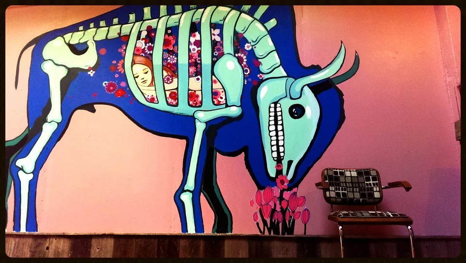 A colorful mural of a blue and green animal, resembling a mix between a horse and a skeleton, with a person resting inside its ribcage, surrounded by flowers, on a pink wall with a patterned chair in front.