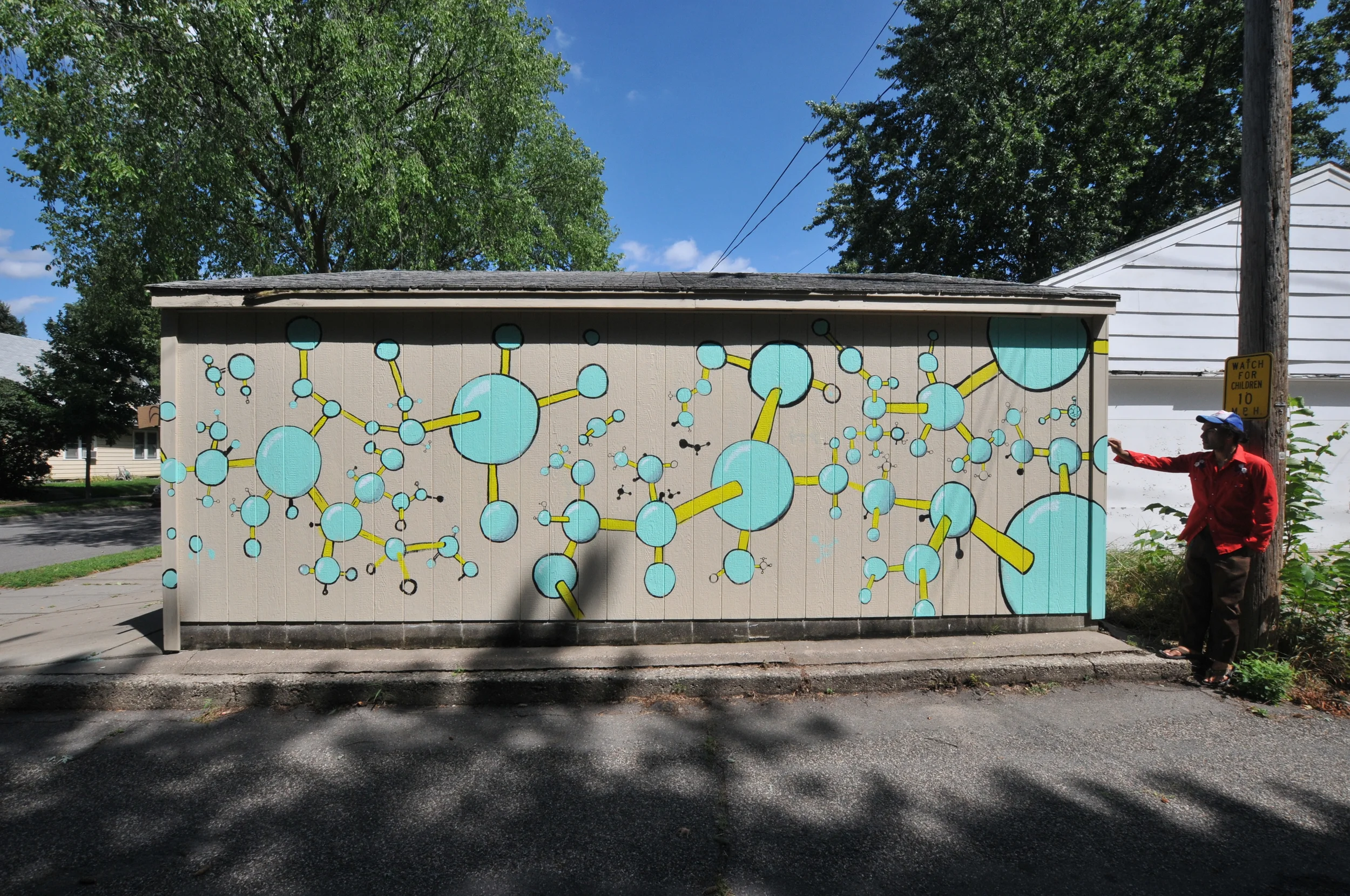 Mural of blue and yellow molecular structures painted on a beige shed, with a person standing to the right pointing at it and a tree and houses in the background.