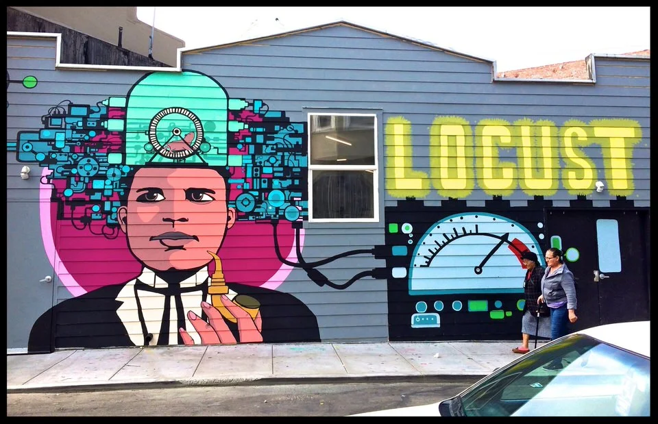 Street art mural depicting a woman with blue circuit-like hair holding a banana, with the word "LO cust" in bright yellow graffiti-style letters, and a speedometer graphic on a building