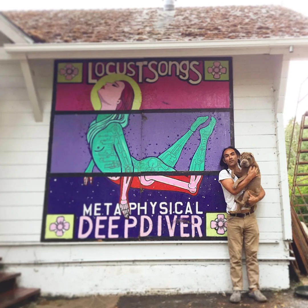A man holding a brown dog standing in front of a colorful mural with a woman lying down and the words "Locust Songs" and "Metaphysical Deep Diver" written on it.