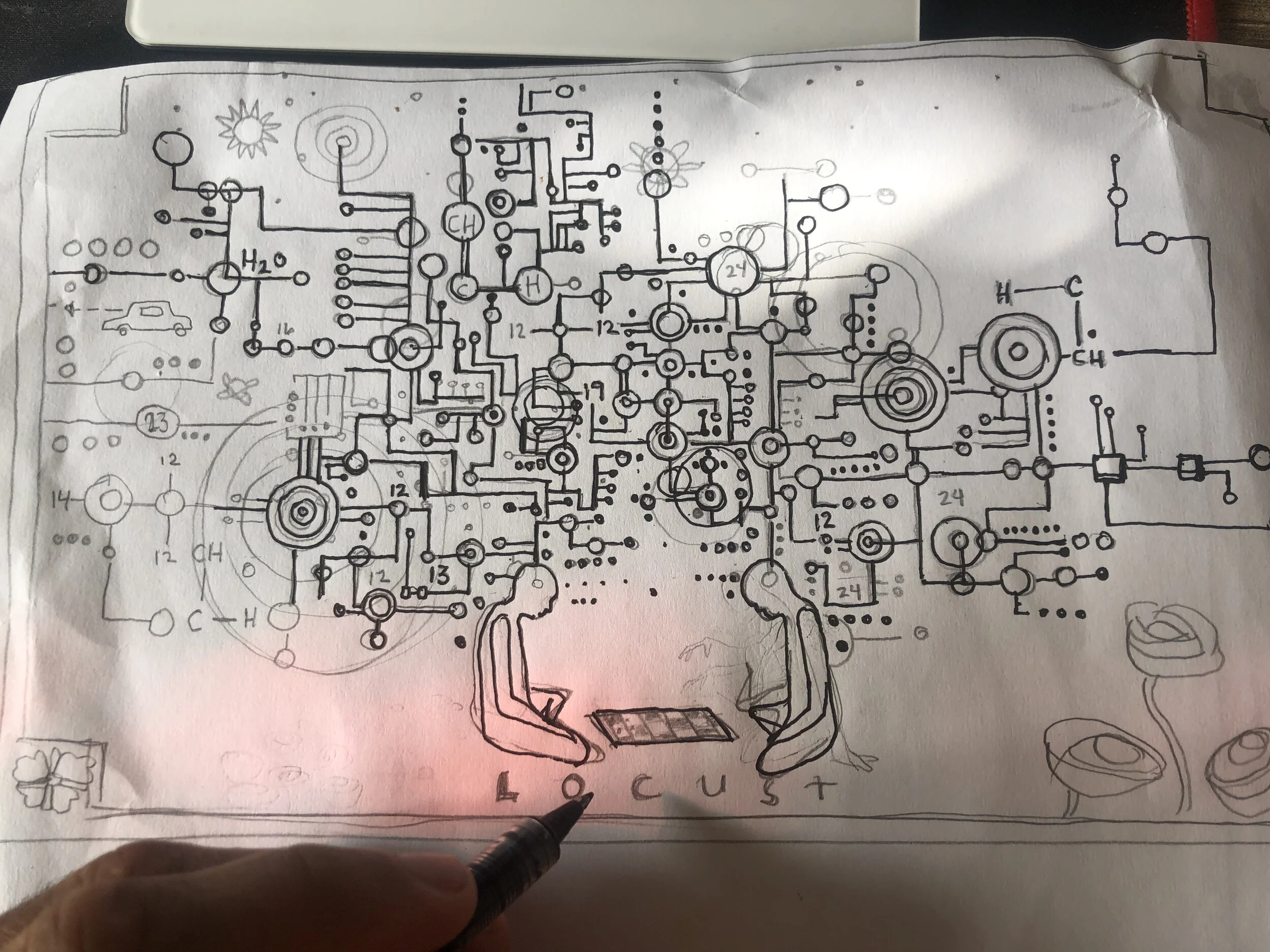 Hand holding a pen pointing at a detailed electronic schematic diagram drawn on paper, showing various circuits, symbols, and annotations.