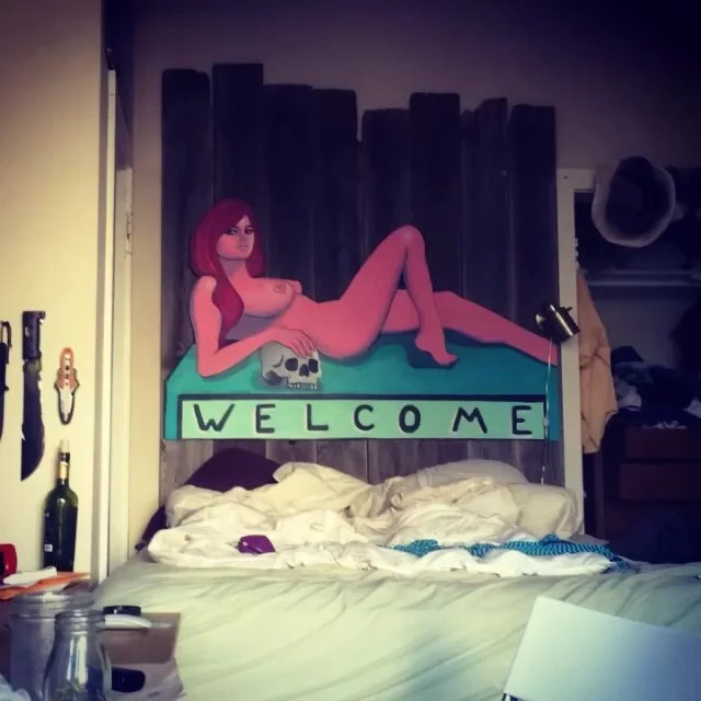 A colorful mural of a woman with pink hair, lying on a bed with a skull, painted above a bed in a bedroom. The mural spells out "WELCOME" with individual letters.