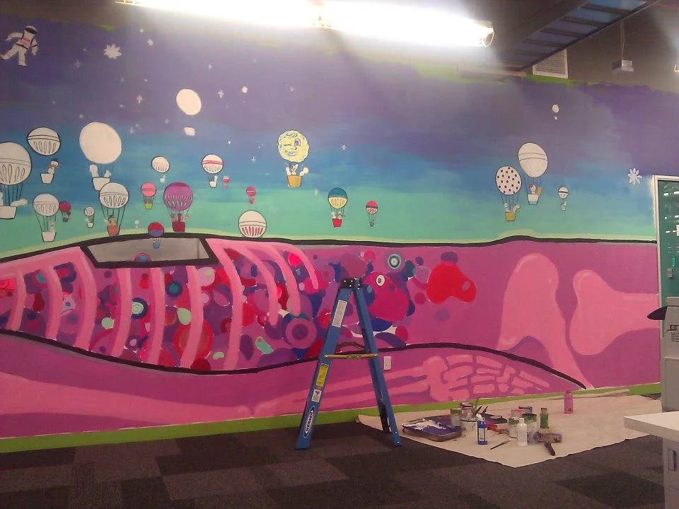 A mural in progress depicting a colorful educational scene with hot air balloons in the sky and a cross-section of a human body showing internal organs, with painting supplies and a ladder in front of it.