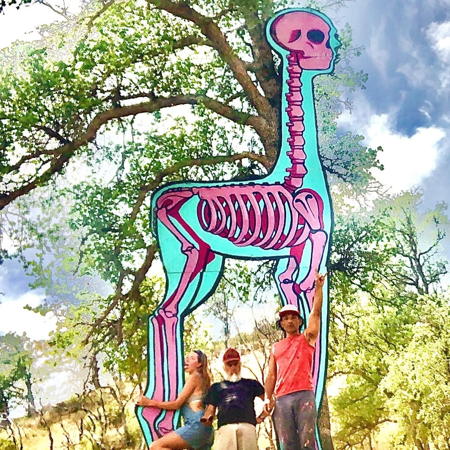 A colorful art installation of a dinosaur skeleton outlined in blue and pink, set outdoors among green trees, with three women standing in front of it, one pointing upward.