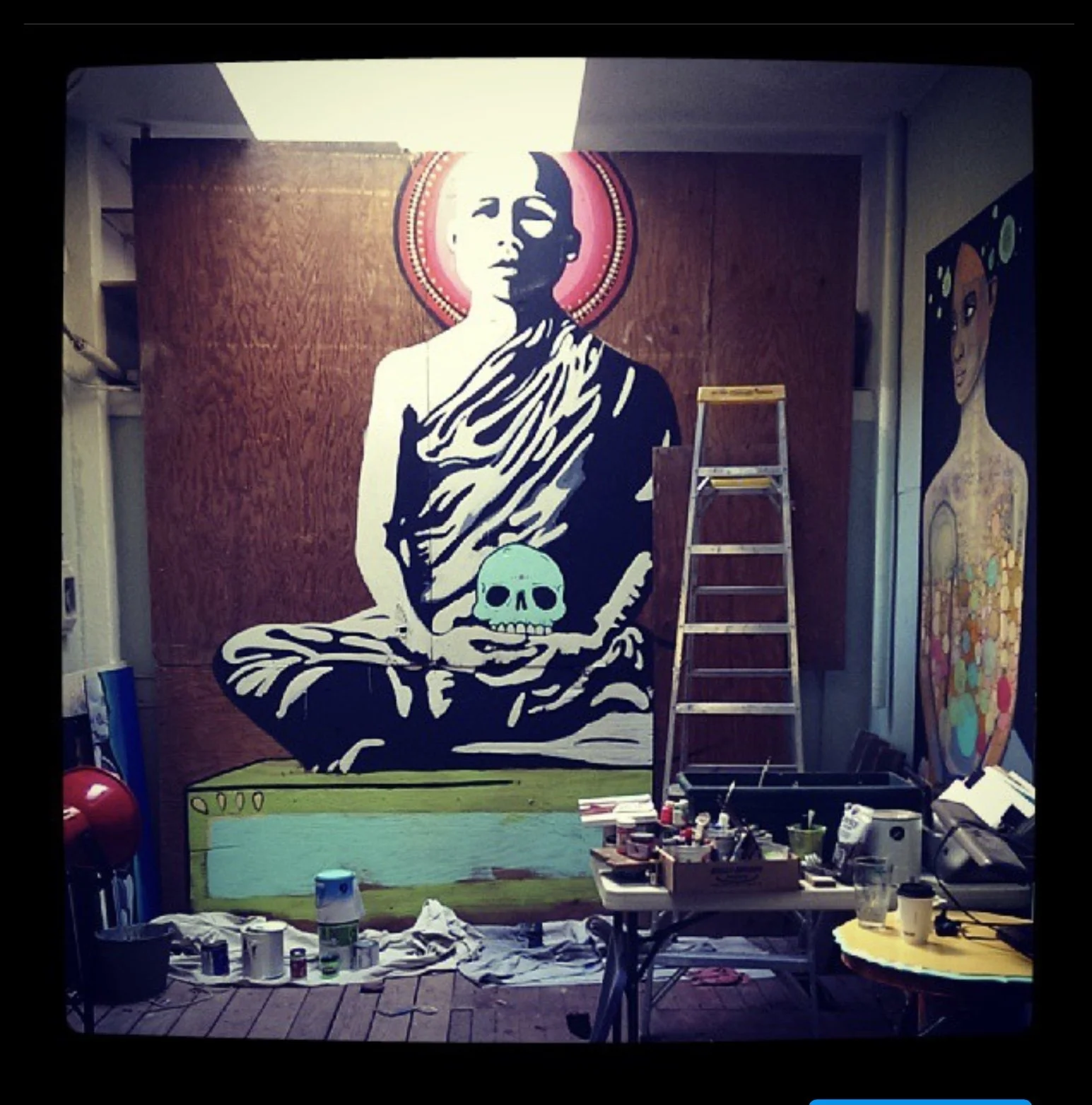 A mural of a Buddha figure holding a skull in front of a brown wooden background, with a red circular halo behind its head. The mural is partially completed, with a ladder in front and art supplies on tables and the floor.