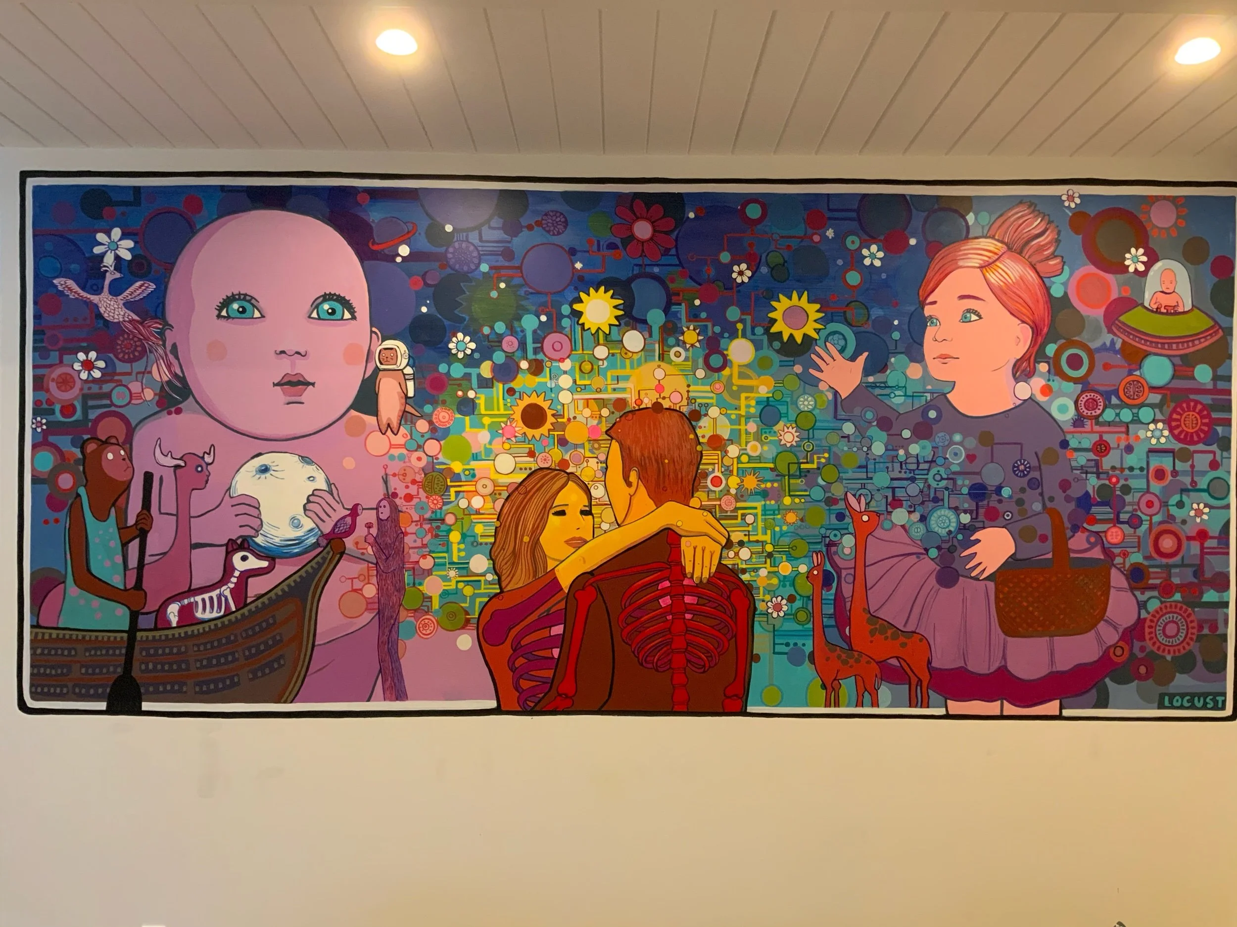 Colorful abstract mural featuring various stylized humanoid figures, animals, and celestial elements, with a large pink baby face on the left, and two children in the center embracing, surrounded by stars, trees, and animals.