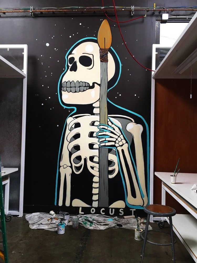 A mural of a skeleton holding a paintbrush with a lit tip, painted on a black wall. The skeleton has a cartoon style with hollow eye sockets and a visible ribcage. The artist's workspace includes paint supplies and furniture around the mural.