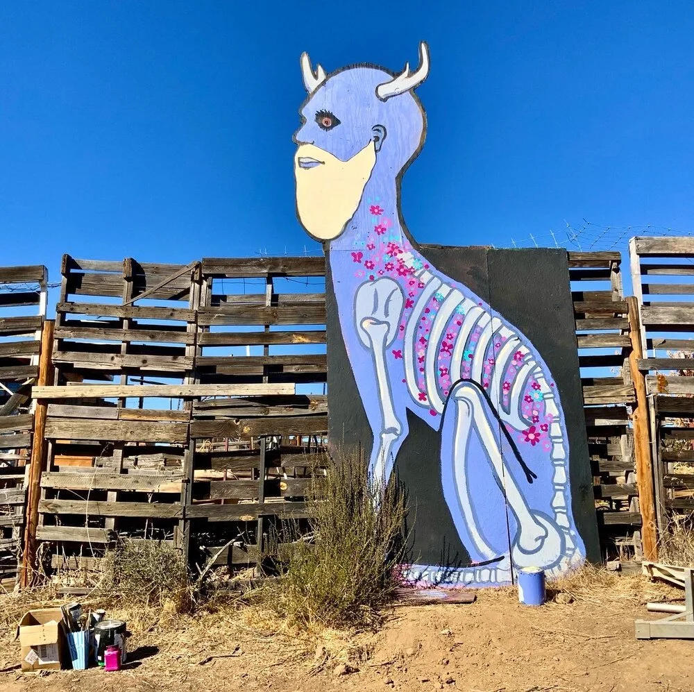 Large painted mural of a hybrid creature with a deer's antlers, a human face with a closed eye, and a body resembling a skeleton with pink flowers on it, against a clear blue sky.