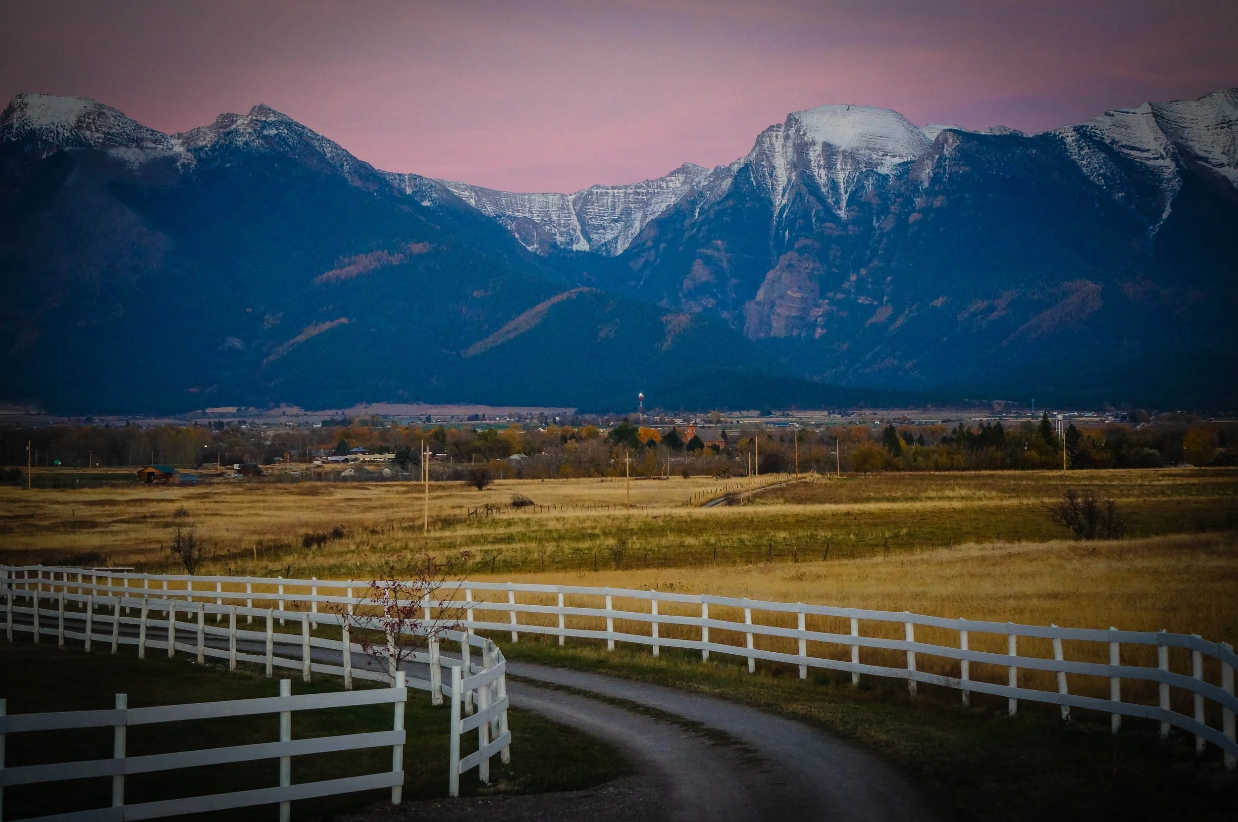 View down the lane at the Rugged Horizon wedding venue in St. Ignatius, Montana, with the snow-capped Mission Mountains in the background. An iconic white fence leads to the venue and the location is surrounded by rolling farmland.